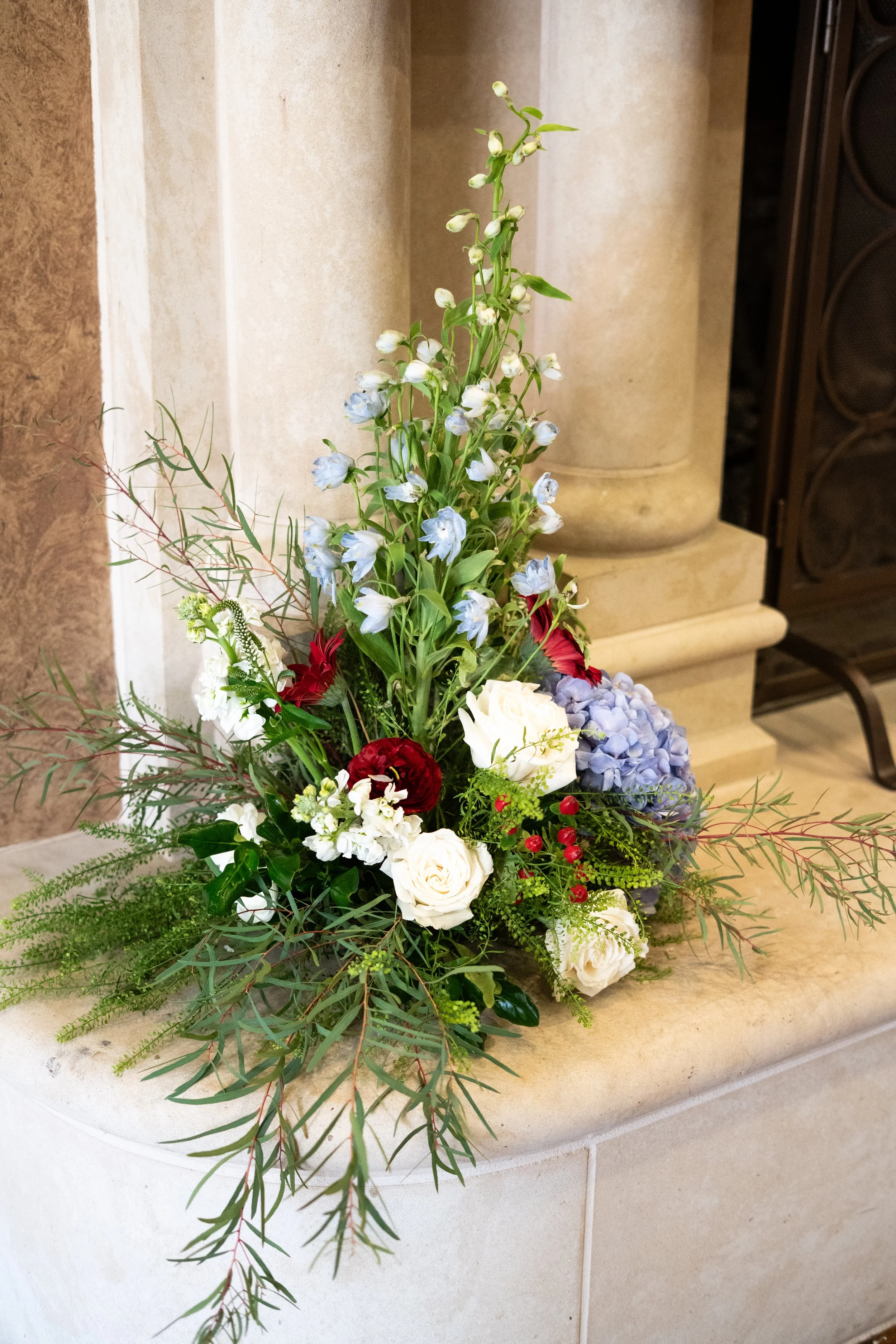 A floral arrangement with white roses, red flowers, and greenery, placed on a stone surface in front of a beige stone column.
