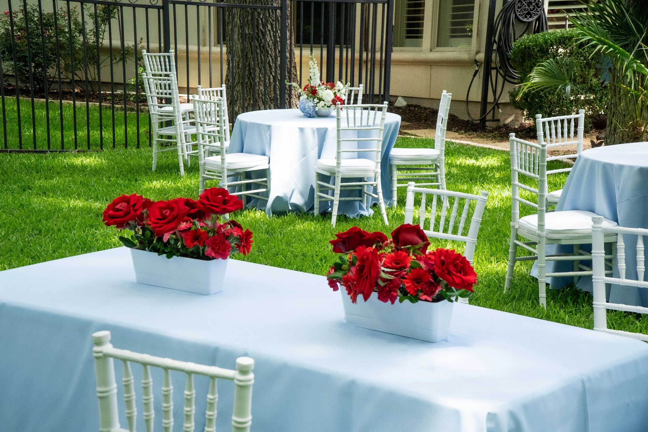 Outdoor event setup with round tables covered with blue tablecloths, white Chiavari chairs, and floral centerpieces with white and red flowers; surrounded by a green lawn, trees, and house exterior.