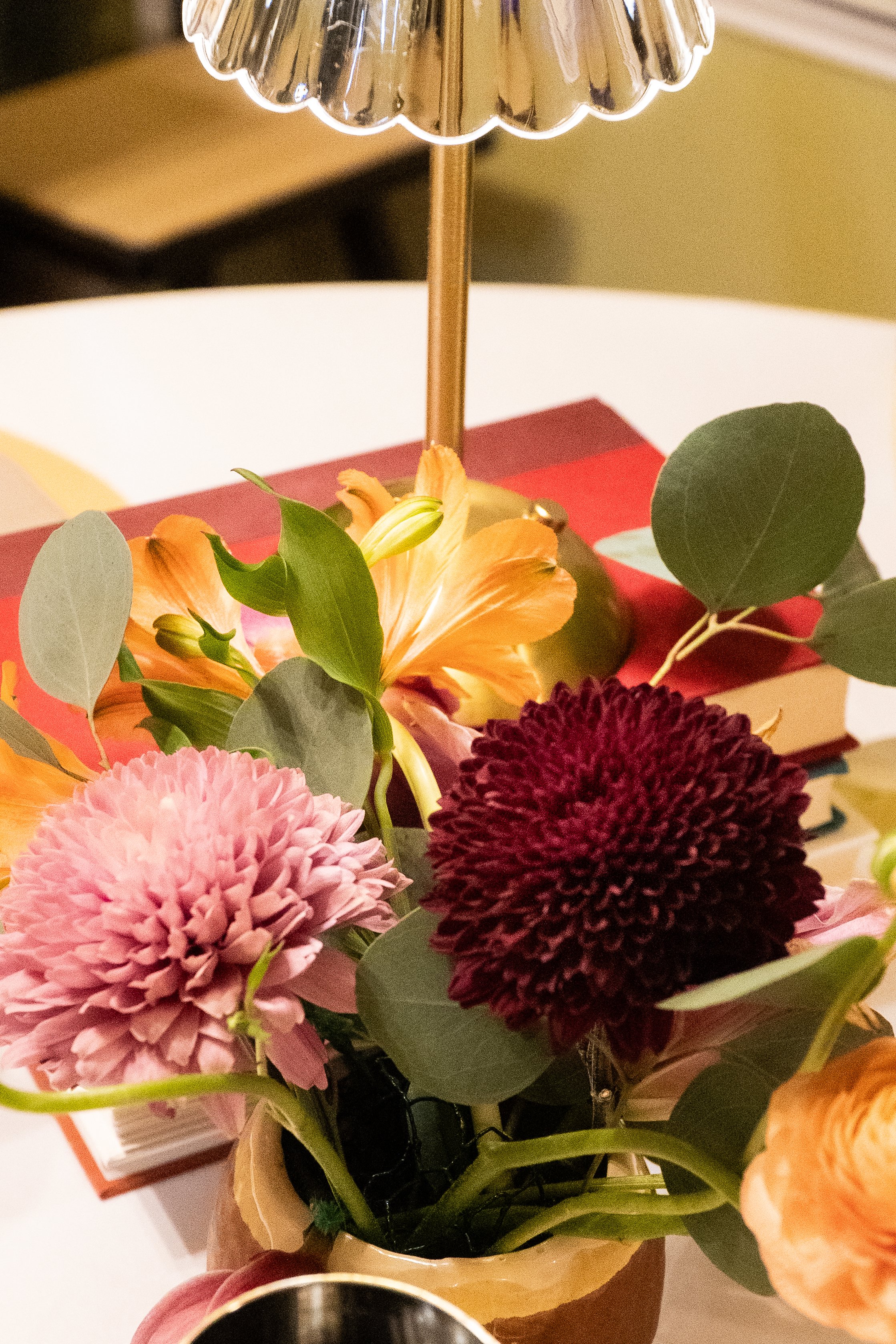 A floral centerpiece with pink, burgundy, and orange flowers, including a pink chrysanthemum and an orange lily, with green leaves, under a shiny, silver lampshade on a table.