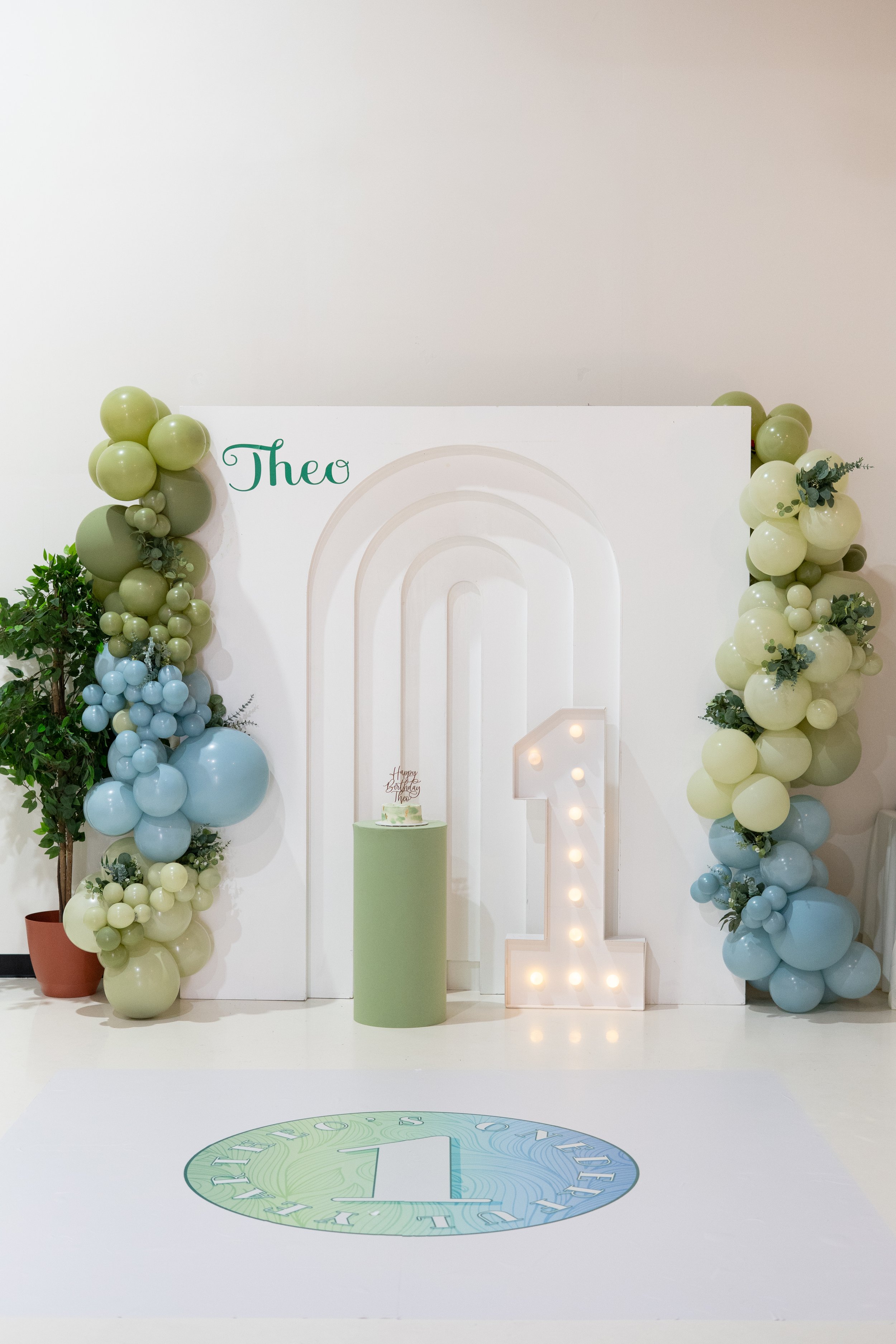 Decorative backdrop with balloons in shades of green and blue, a white panel with the name 'Theo' in green, and a large illuminated number '1' for a first birthday celebration.