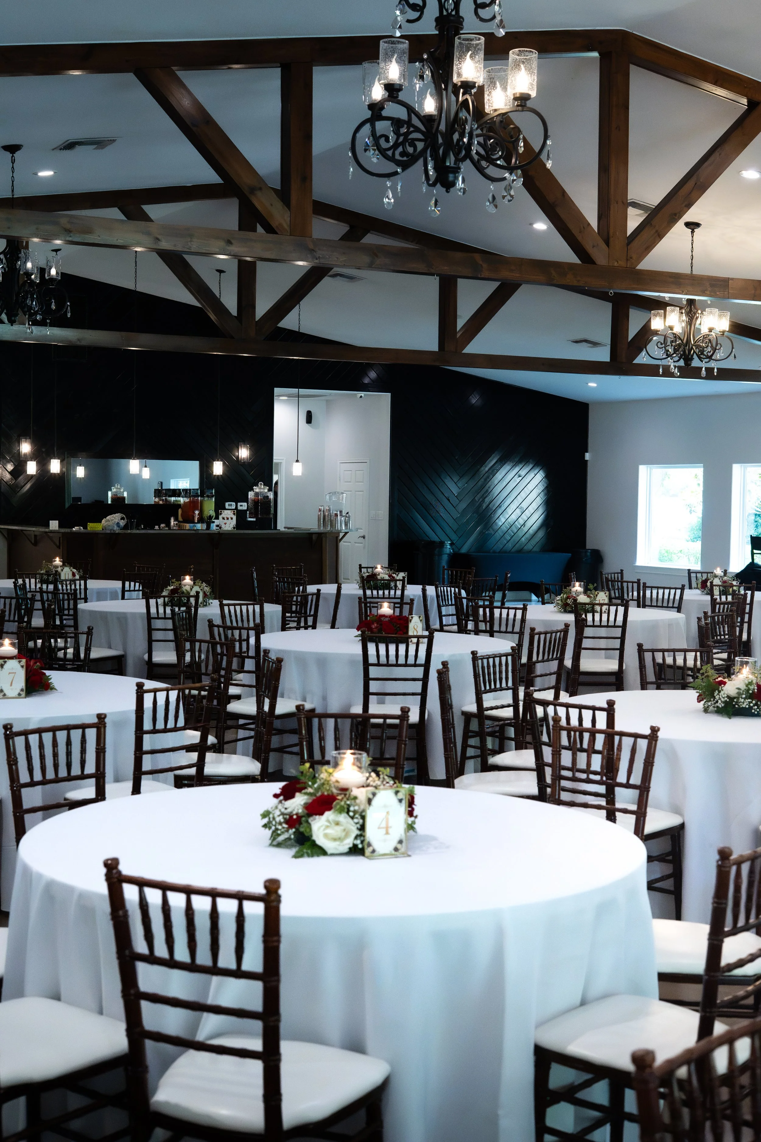 Indoor event space with round tables covered in white tablecloths, decorated with floral centerpieces, surrounded by dark wooden chairs. The ceiling features exposed wooden beams and ornate chandeliers. In the background, a bar area is visible with l