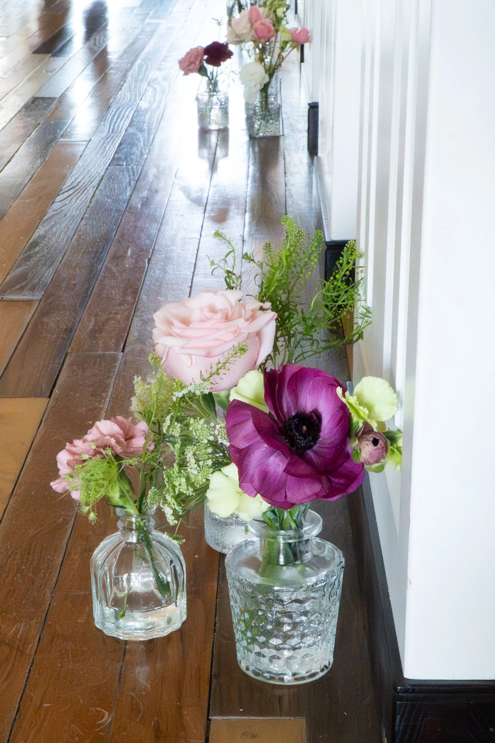 A row of glass vases filled with pink, purple, and white flowers placed on a wooden floor along a white wall. Dallas Florist, wedding flowers north Texas.