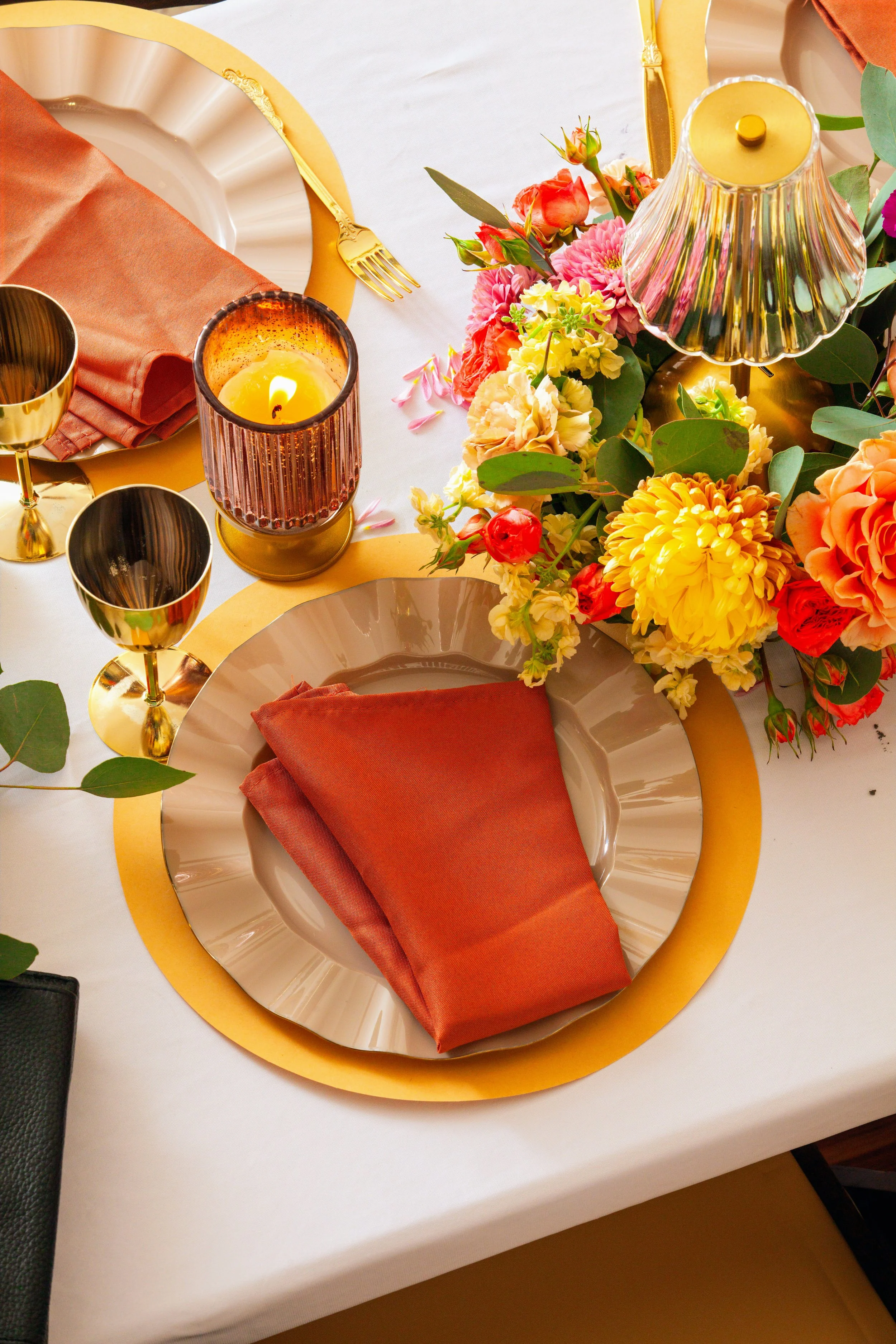A table setting with a gold and white plate, a rust-colored napkin, a gold goblet, a lit candle in a pink glass holder, and a floral centerpiece with colorful flowers including pink, yellow, and orange blooms on a white tablecloth.