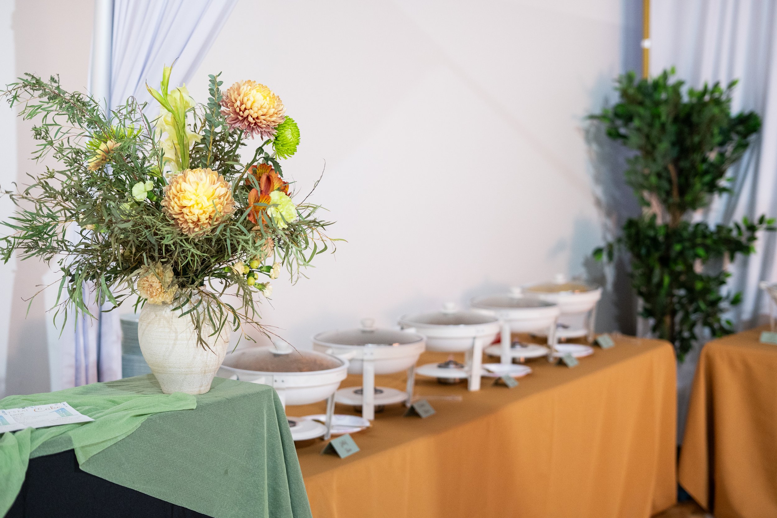 A white vase with a bouquet of mixed flowers including dahlias, green foliage, and smaller flowers on a green tablecloth, with shelves and potted plants in the background.