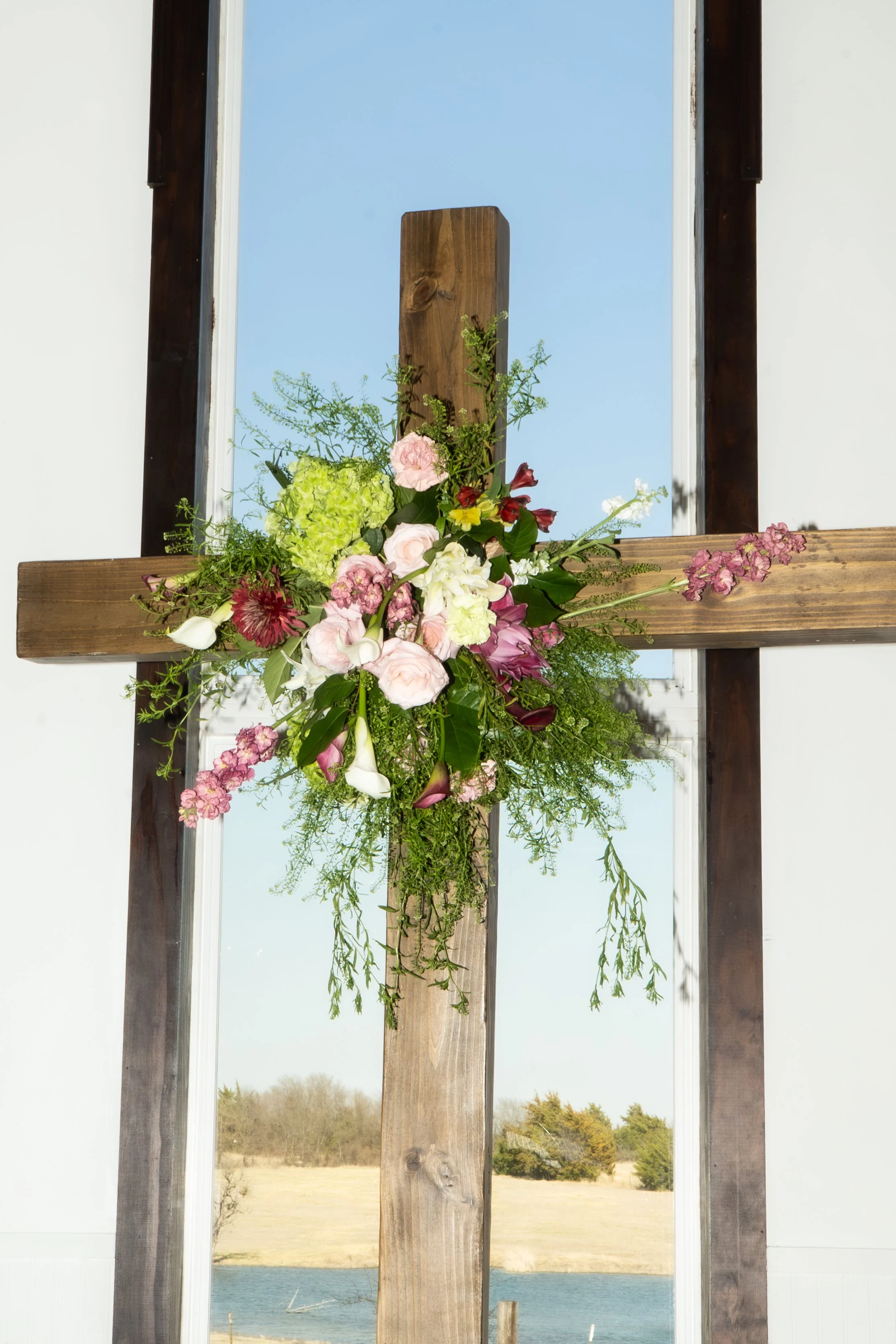 A wooden cross decorated with a floral arrangement, positioned in front of a large window with a blue sky and outdoor landscape visible in the background.