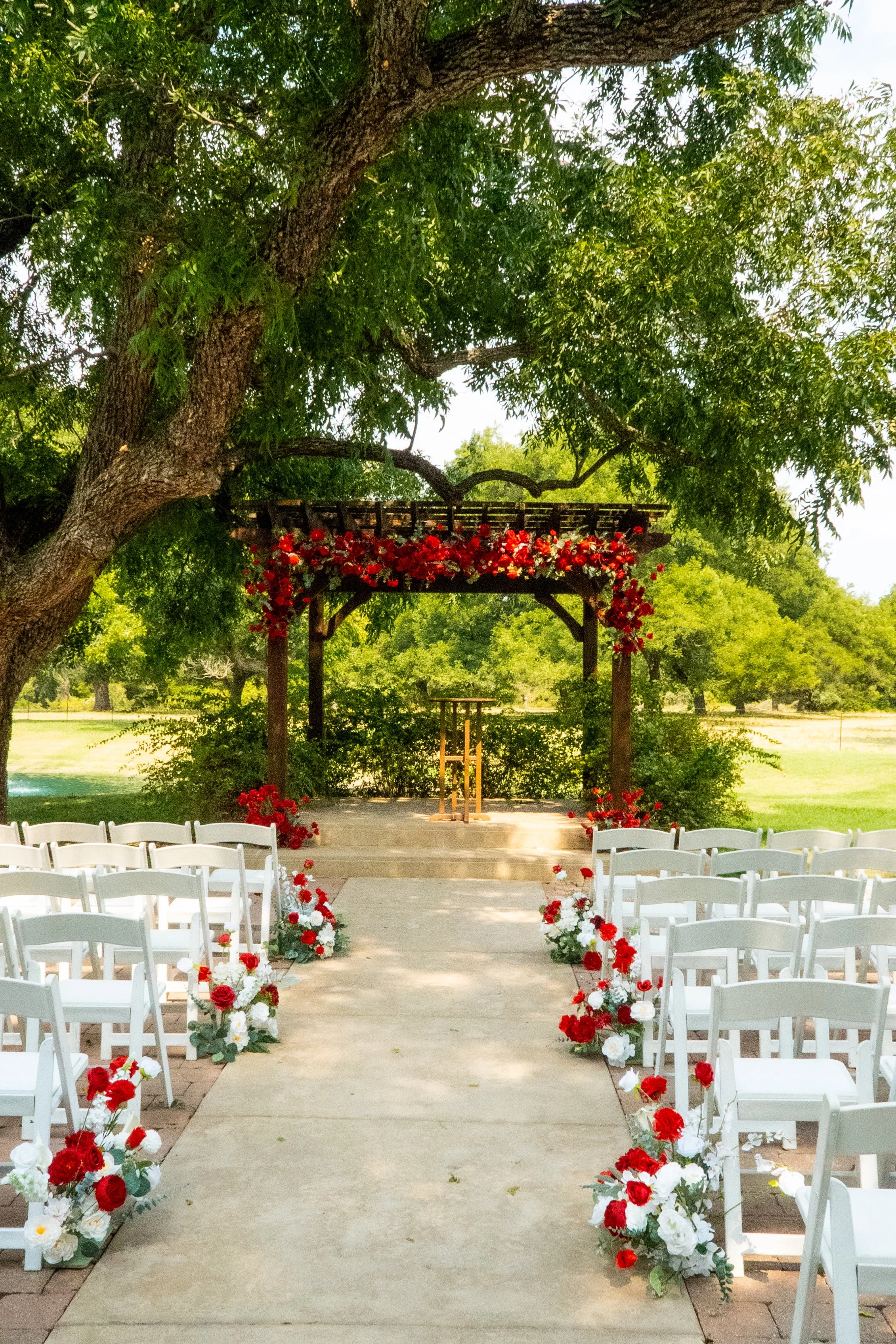 Outdoor wedding ceremony setup with white chairs lining a central aisle decorated with red and white floral arrangements, under a wooden arbor draped with red flowers, in a park-like setting with green trees and grass.