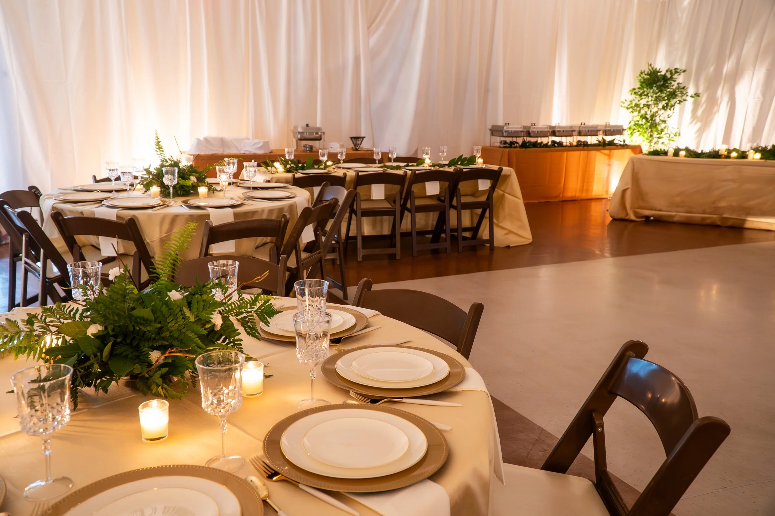 Restaurant banquet setup with round tables, white tablecloths, candle centerpieces, and chairs, decorated with greenery, and buffet tables against a cream-colored curtain backdrop.