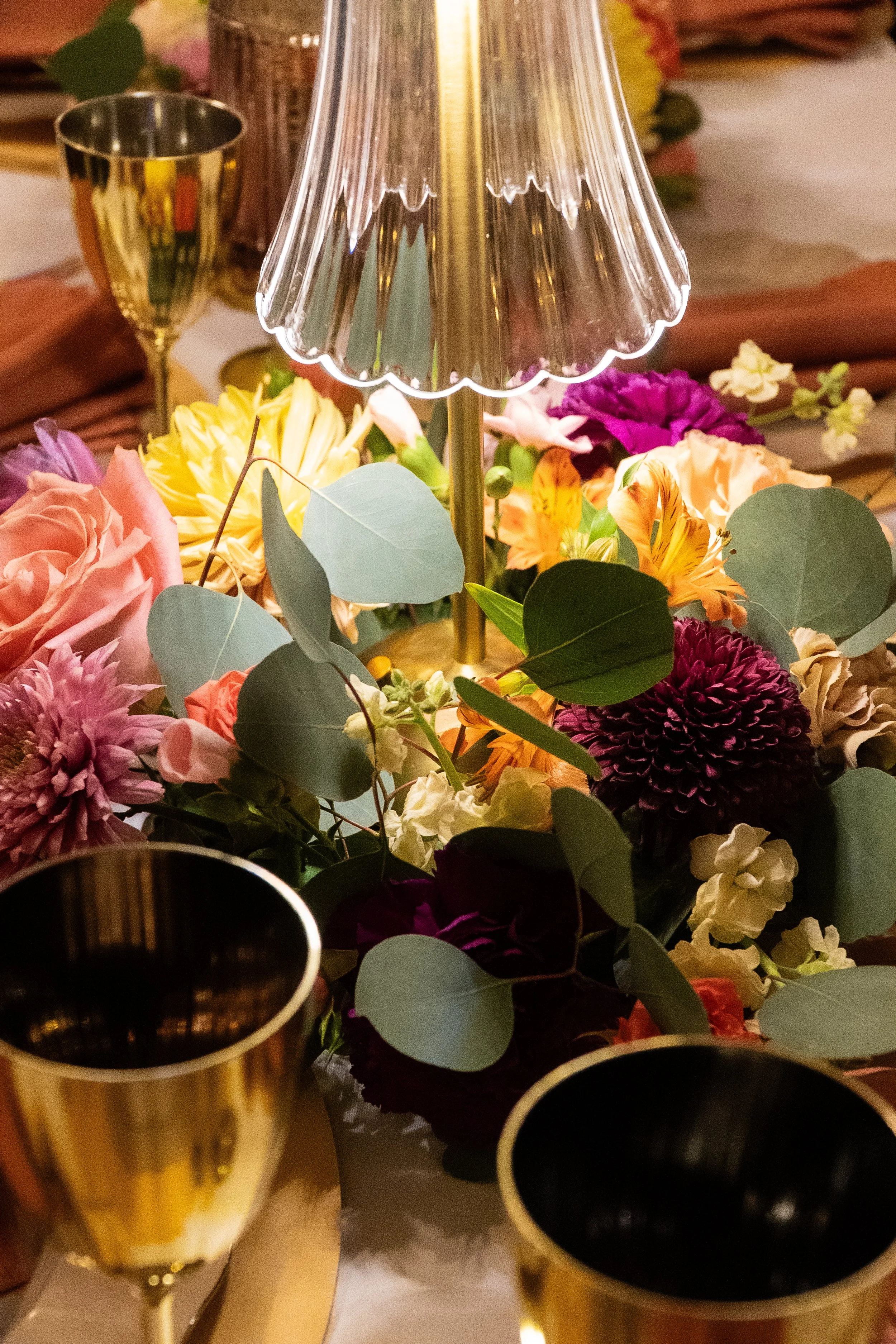 Close-up of a table centerpiece with colorful flowers, eucalyptus leaves, a glass lamp in the center, and gold-rimmed wine glasses surrounding it.