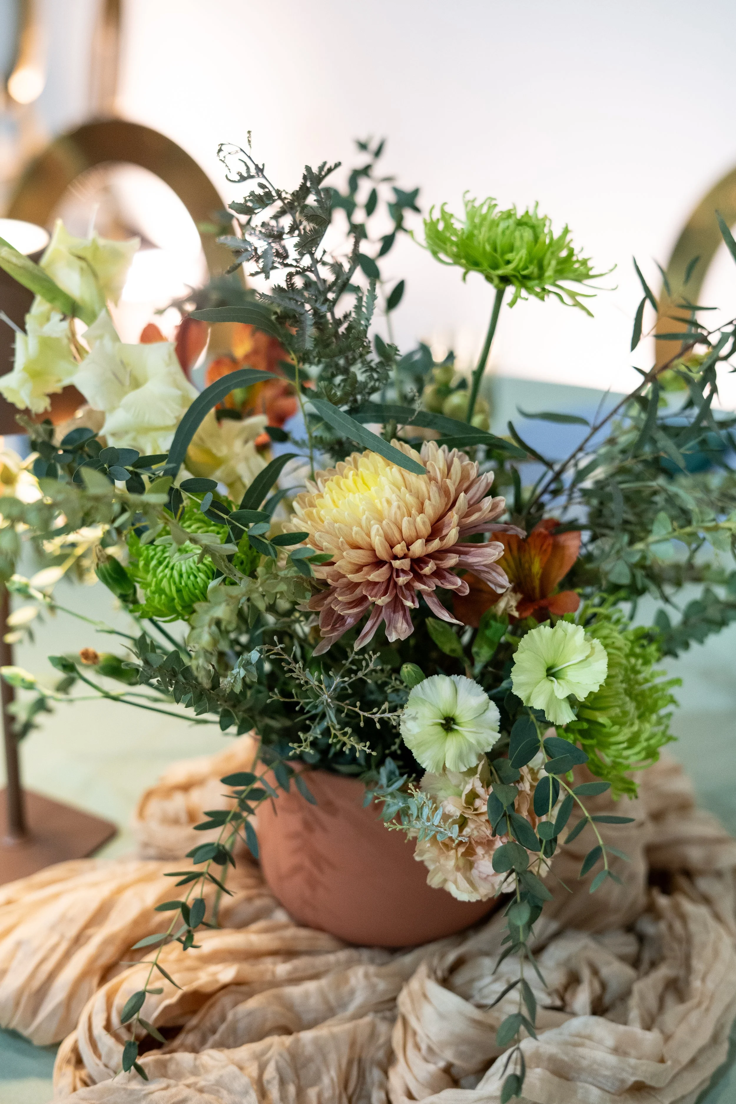 A floral arrangement with a variety of flowers and greenery in a brown ceramic pot on a textured beige surface.