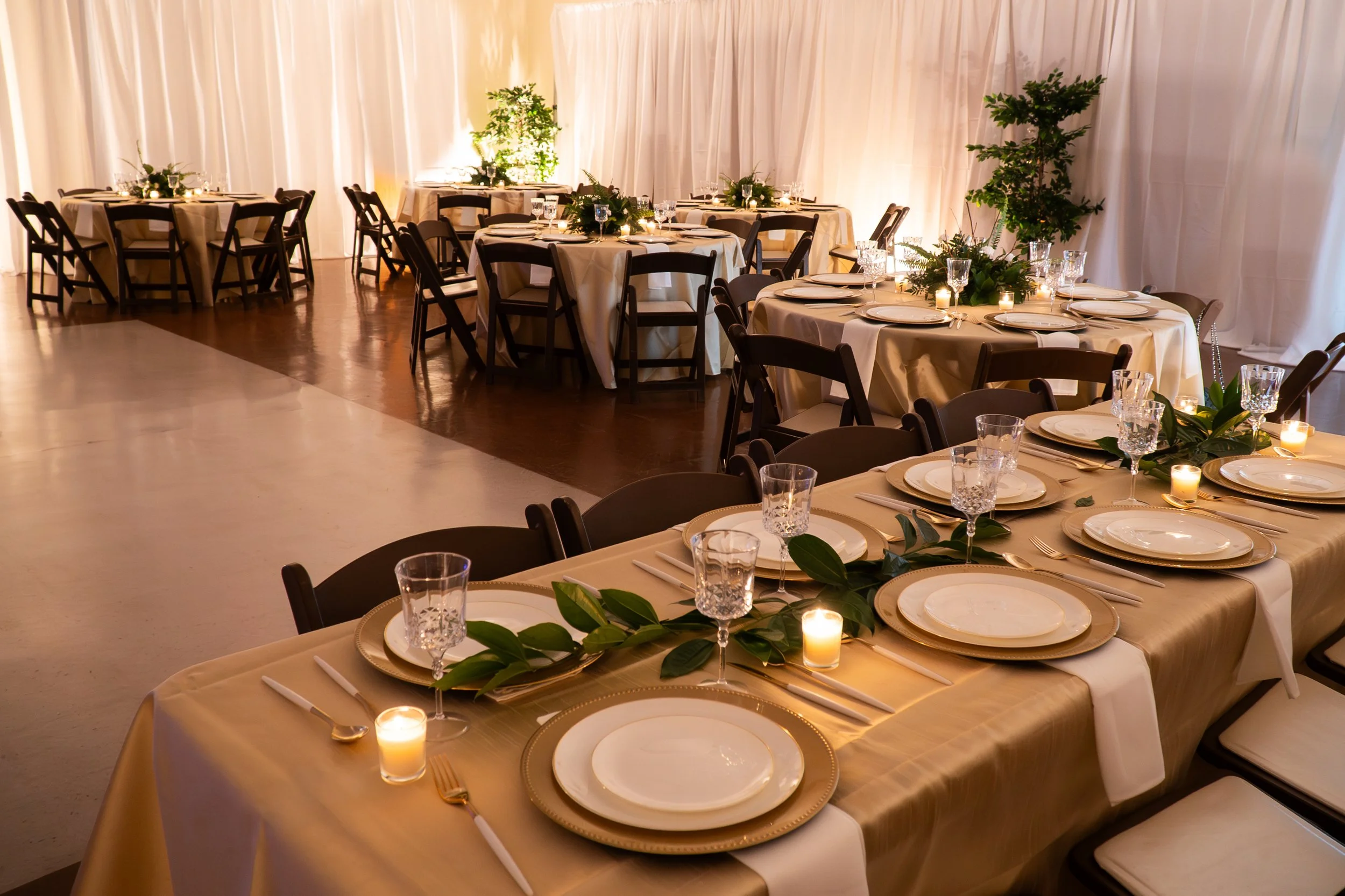Elegant dining setup with round tables covered in beige tablecloths, set with white plates, glassware, gold utensils, and green foliage centerpieces illuminated by small candles, with cream-colored curtains in the background.