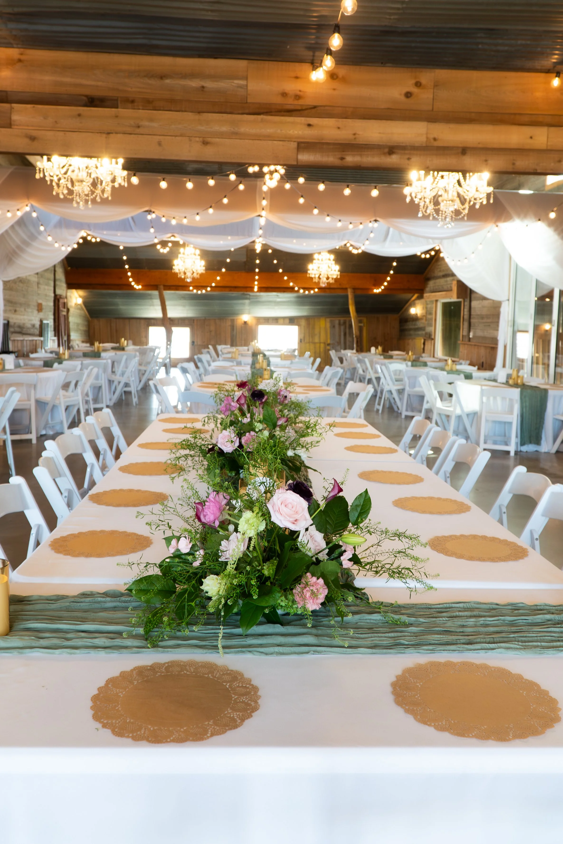 Wedding reception hall with long tables, floral centerpiece, white chairs, string lights, chandeliers, wooden walls, and draped fabric ceiling.