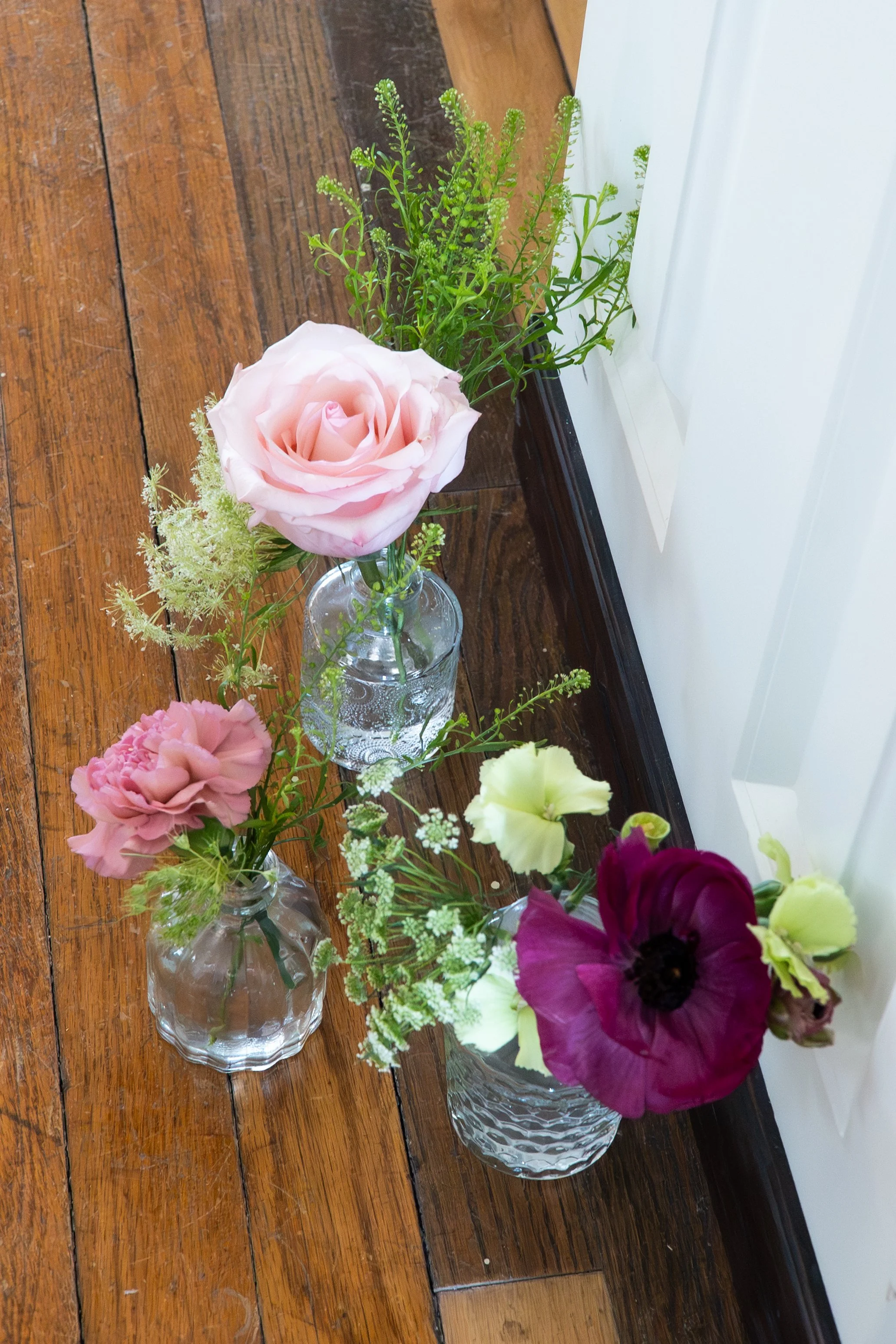 Three small glass vases with mixed flowers, including roses and anemones, placed on a hardwood floor near a window.