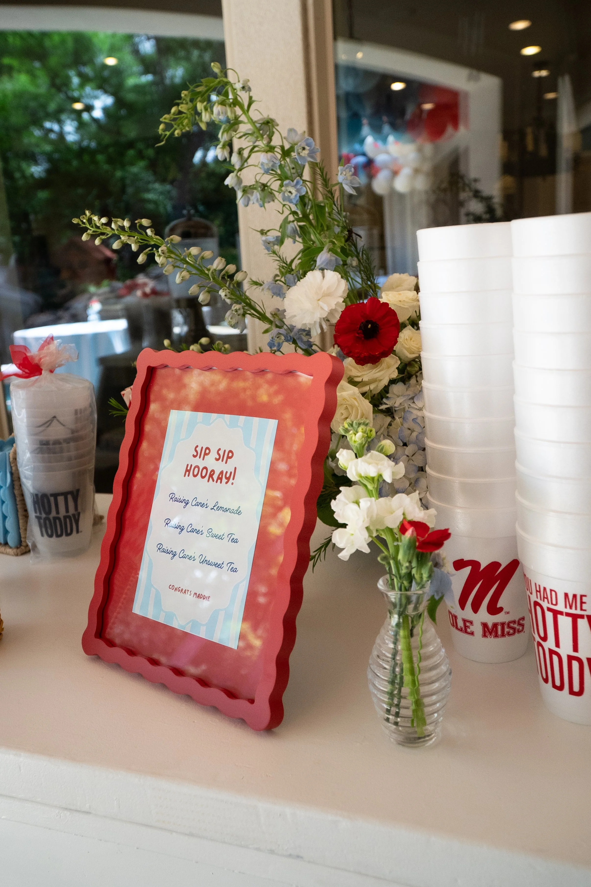 A small glass vase with a bouquet of white, red, and light blue flowers on a white countertop. There is a framed sign with a pink scalloped border and blue striped background that reads "Sip Sip Hooray!" and a list of beverages, with a message "Congr