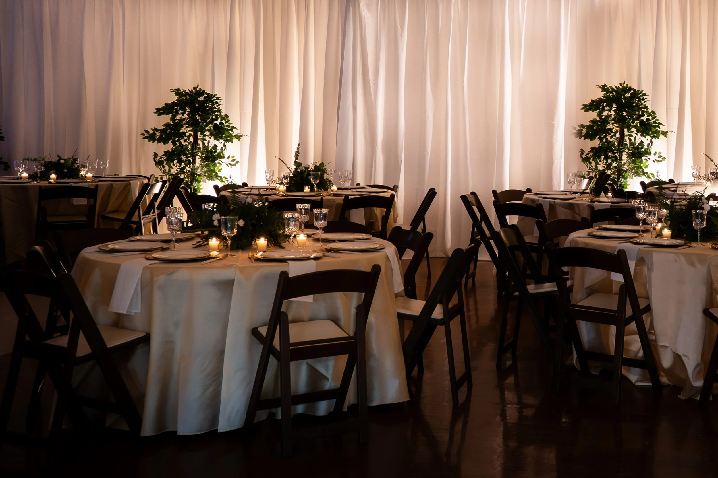 Elegant dining setup with round tables covered in white tablecloths, decorated with candles, glassware, and floral centerpieces, set against a backdrop of white curtains.