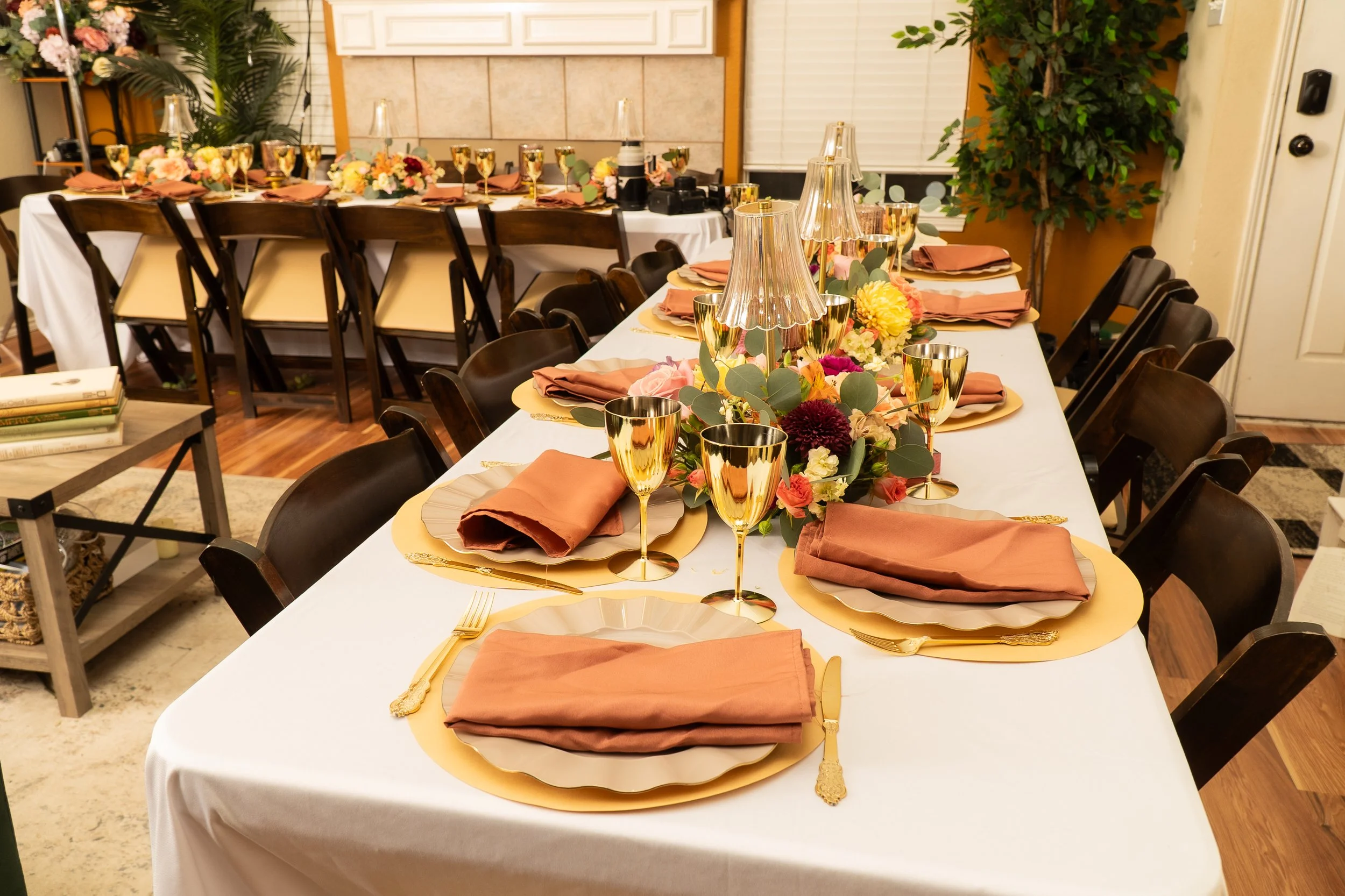 A dining table set for a formal event with white tablecloth, gold chargers, pink napkins, gold cups, and a floral centerpiece with pink, yellow, and burgundy flowers, surrounded by wooden chairs. Texas florist, ft worth wedding flowers  wedding