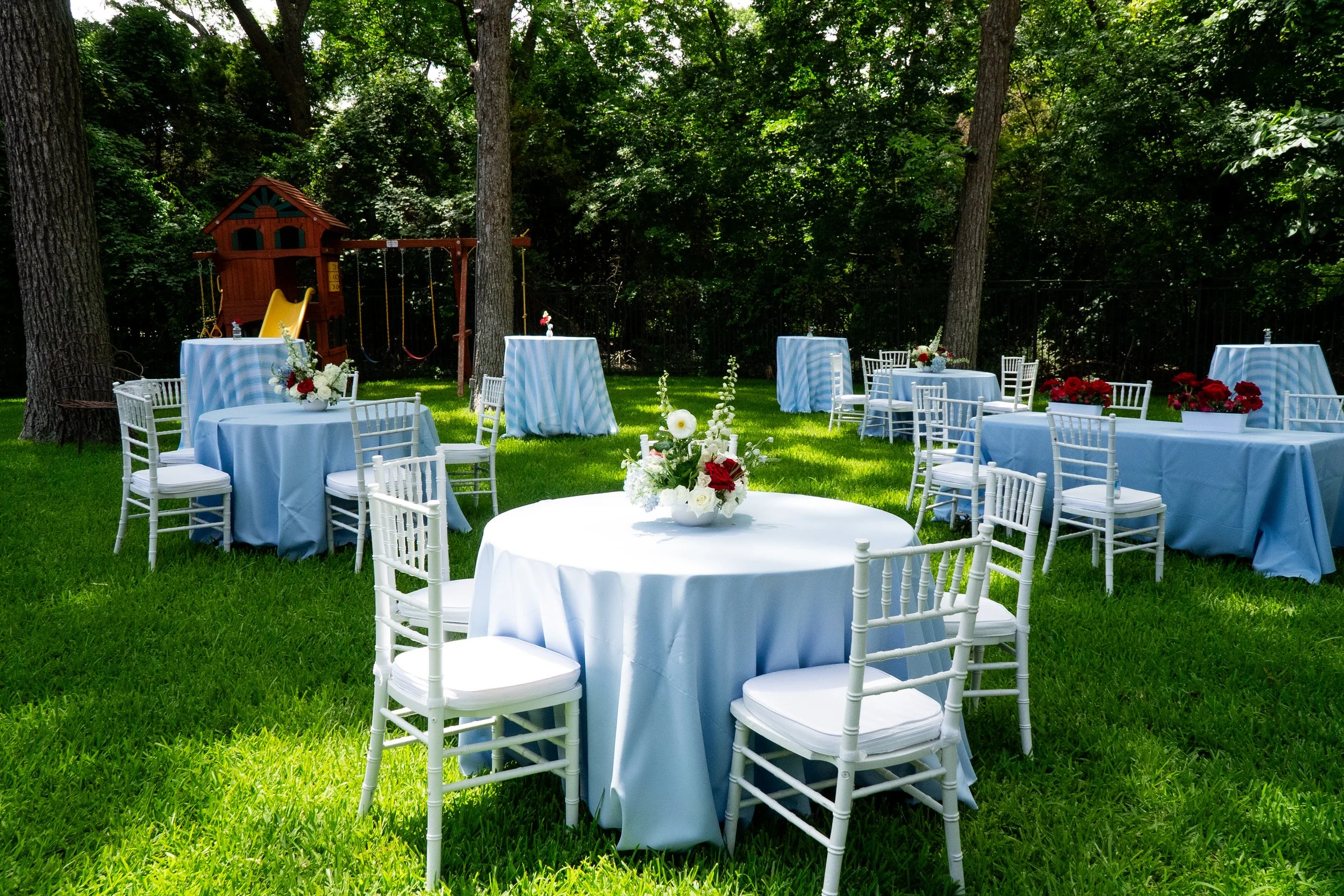 Outdoor garden decorated with white tables covered in blue and white striped tablecloths and white chairs, set for an event with floral centerpieces. There is a children's playset and swing set in the background among tall trees and greenery.