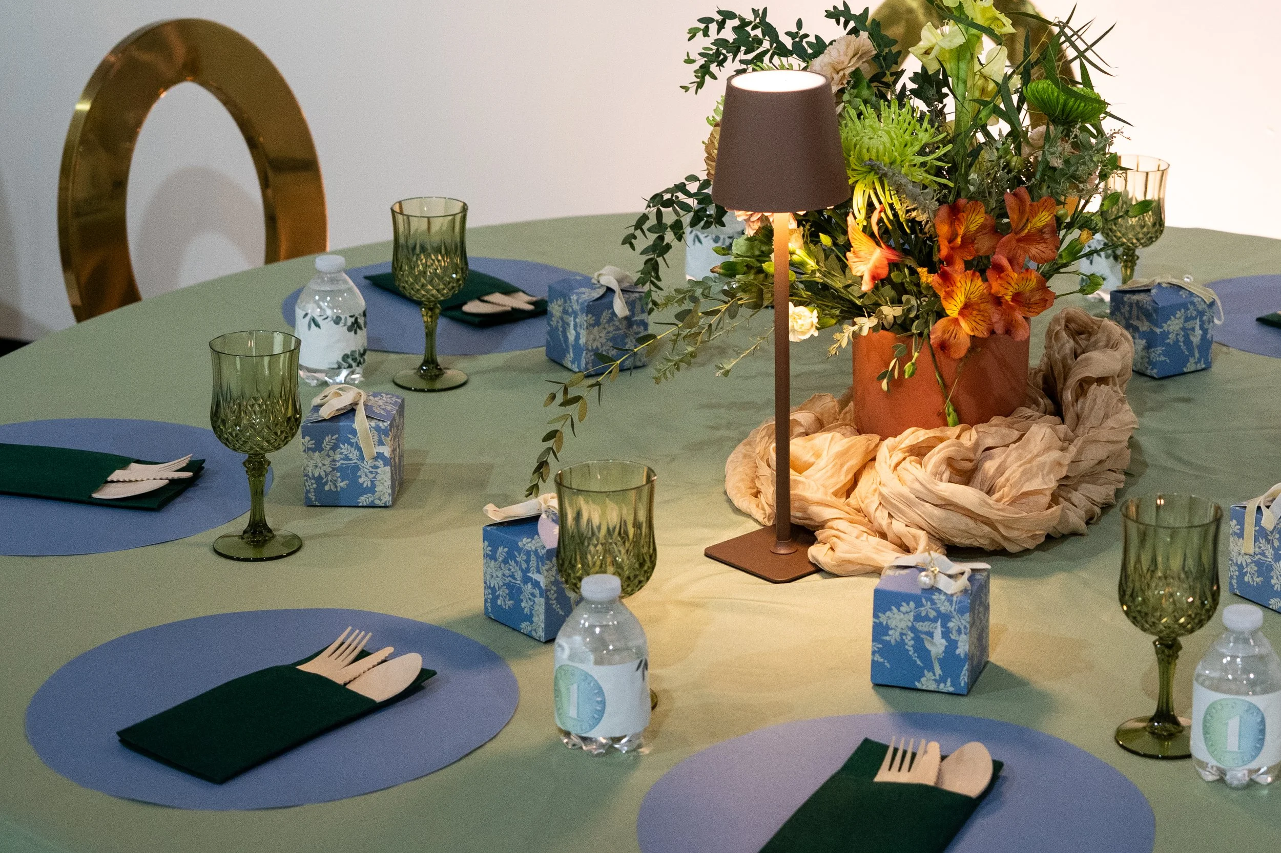 A decorated dining table with a floral centerpiece, tableware, glasses, and presents, illuminated by a table lamp.