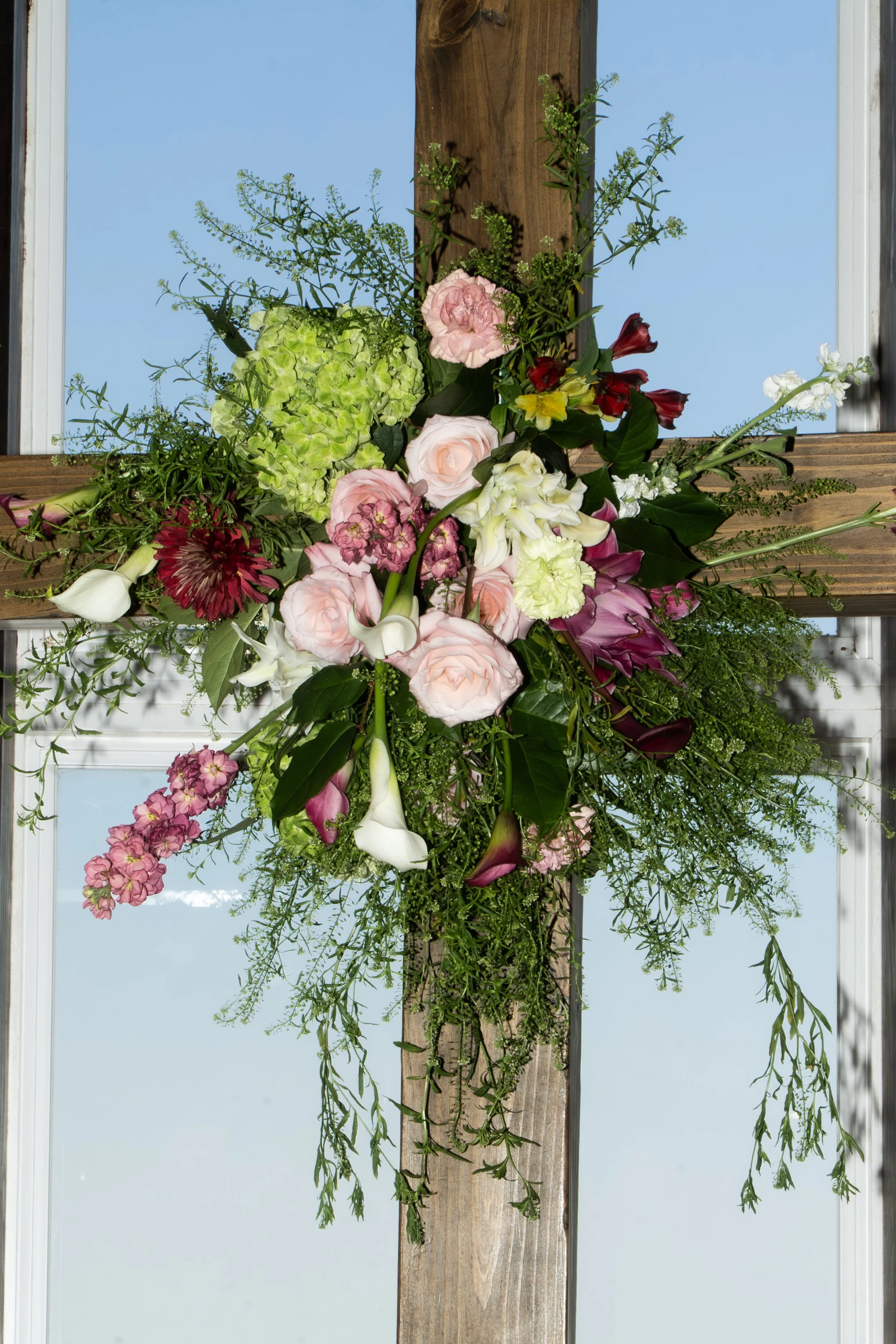 An arrangement of pink, white, purple, and green flowers and greenery mounted on a wooden cross, set against a bright blue sky. fort worth flowers