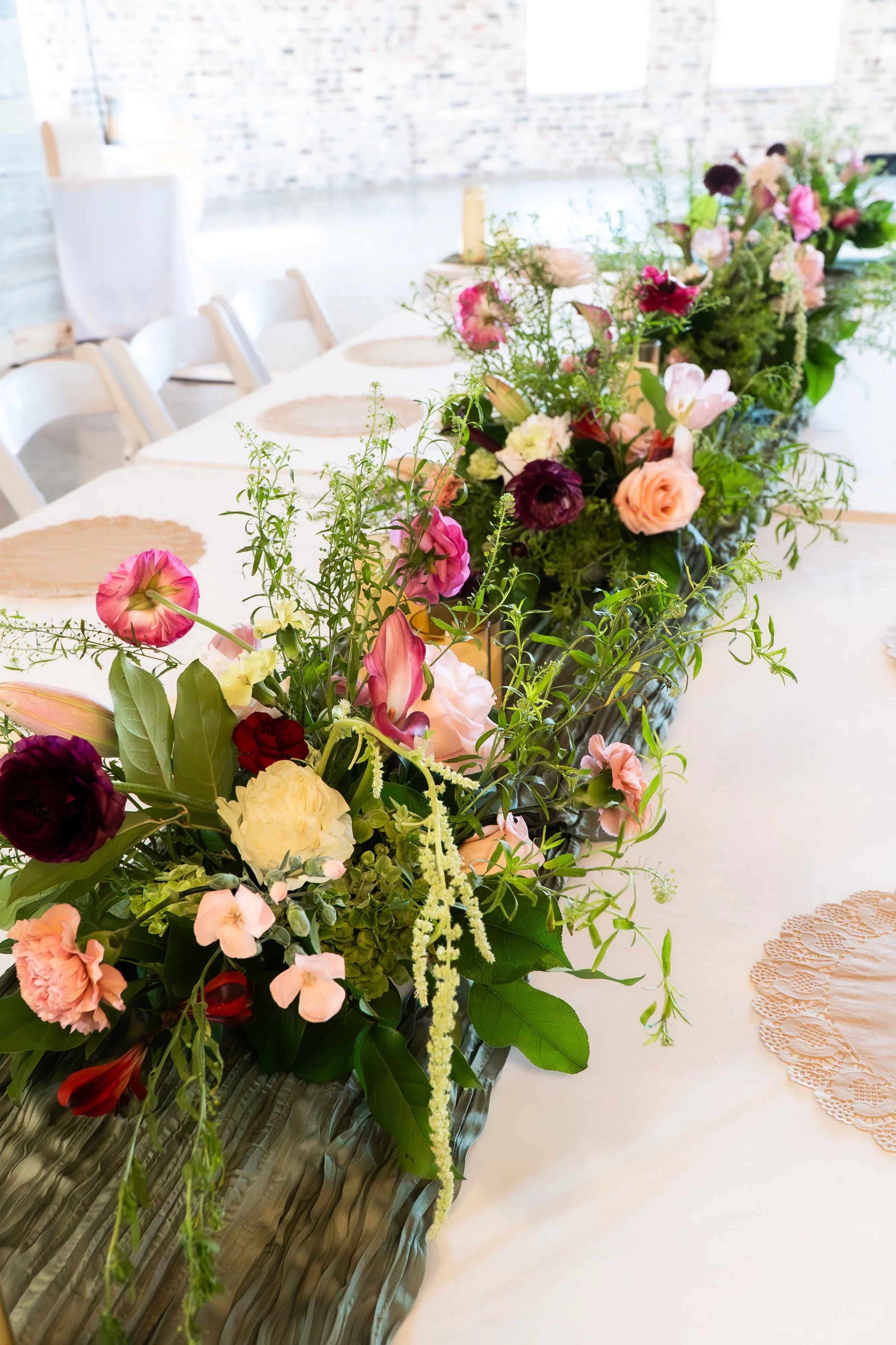 A long table decorated with a floral centerpiece with pink, purple, white, and peach flowers, and paper placemats with doilies in a bright indoor space.