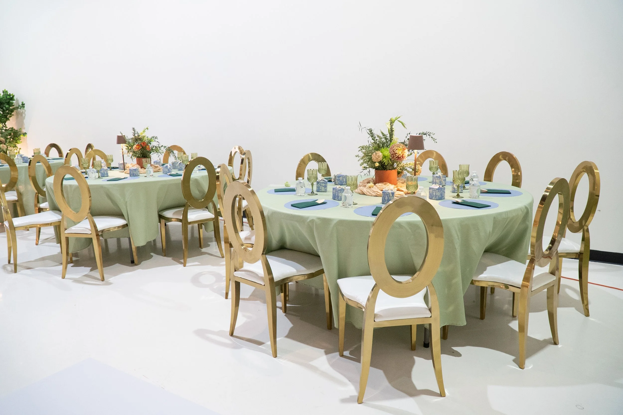 Round banquet table with green tablecloth, decorated with floral centerpieces, set with glasses, napkins, and small boxes, surrounded by chairs at a formal event space.