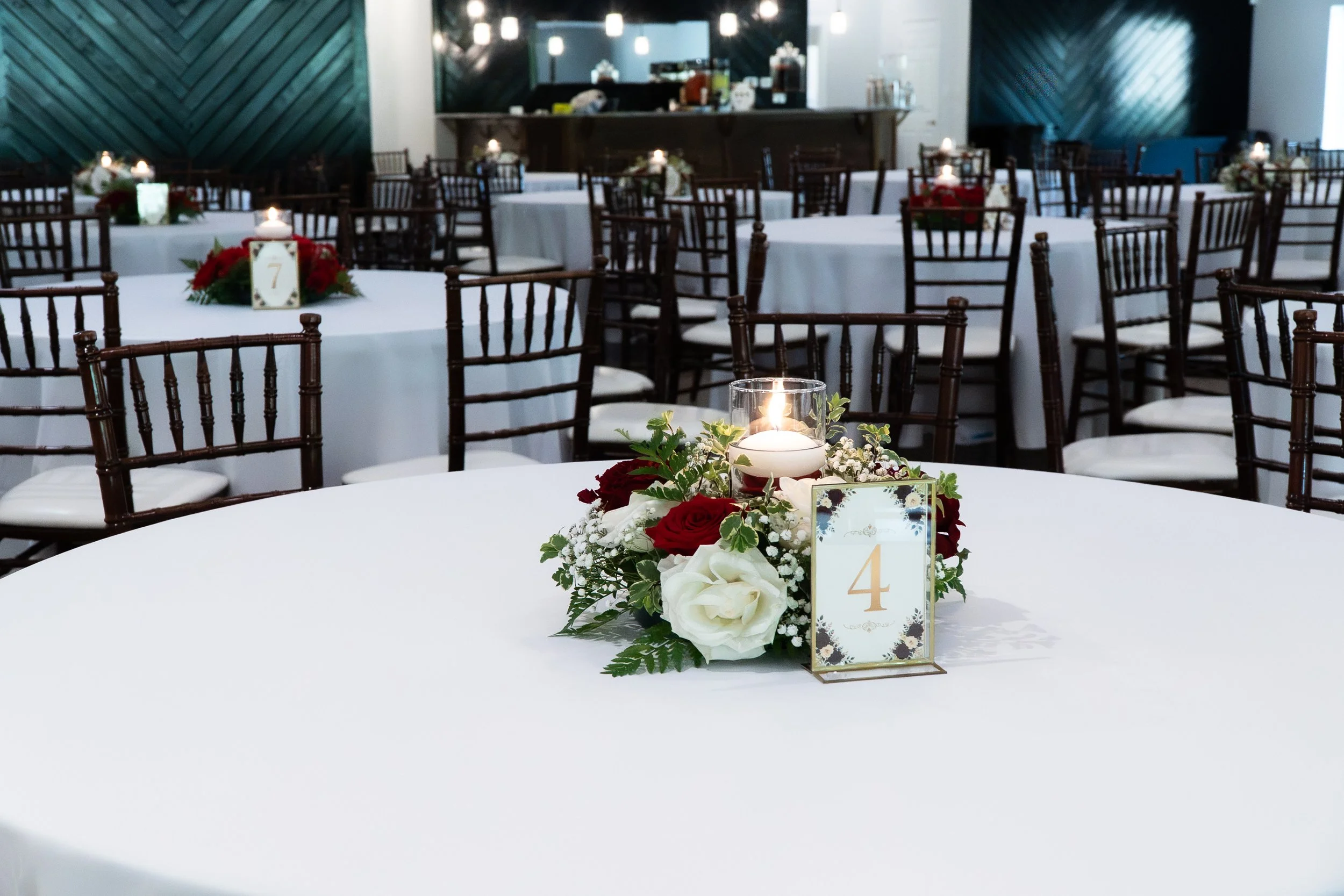 Elegant banquet table decorated with a floral arrangement, candles, and table number, set in formal event setting with additional tables and dim lighting.