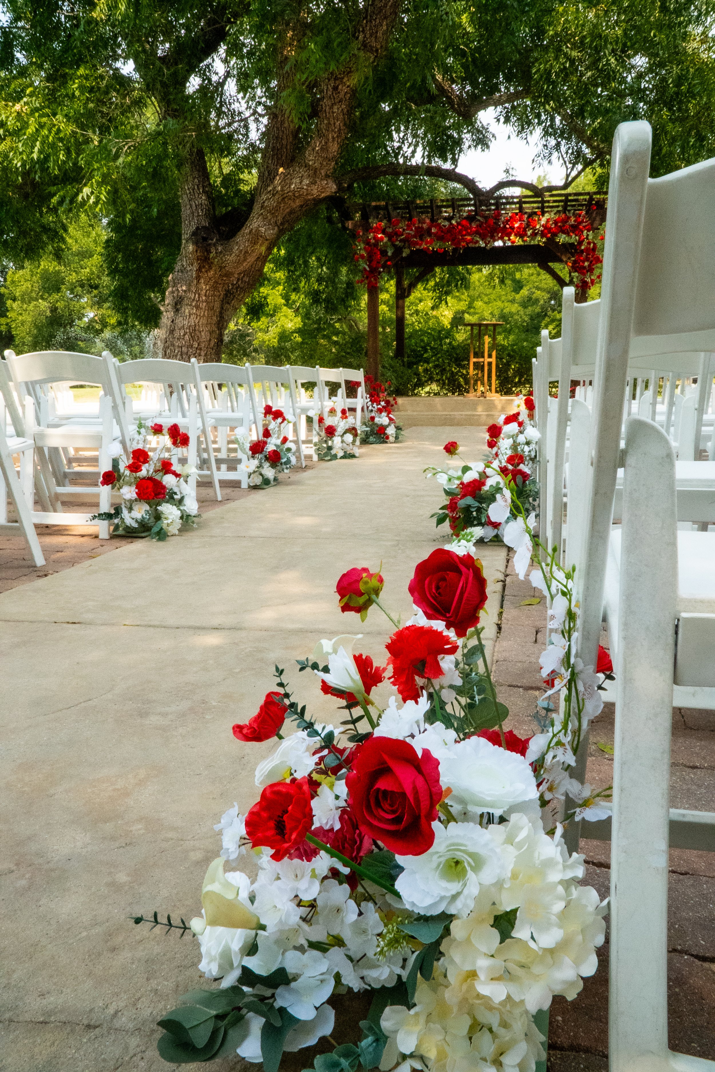 A decorated outdoor ceremony setup with white chairs, red and white floral arrangements, a tree, and a floral arch in the background.