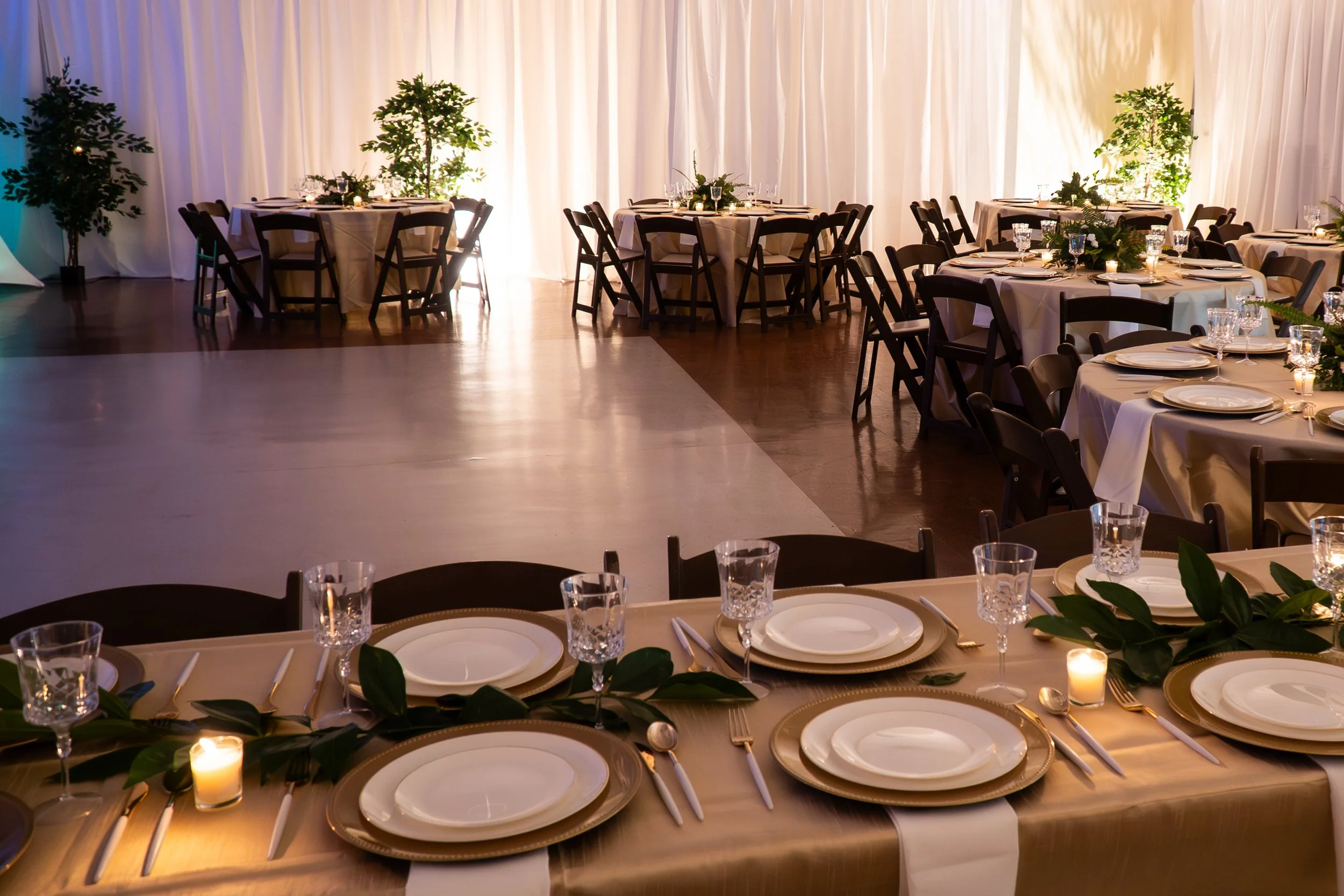A banquet hall decorated for a formal event with round tables covered in white tablecloths, set with plates, silverware, wine glasses, and candles. Green foliage and candles serve as centerpieces, with a backdrop of white curtains and potted plants.