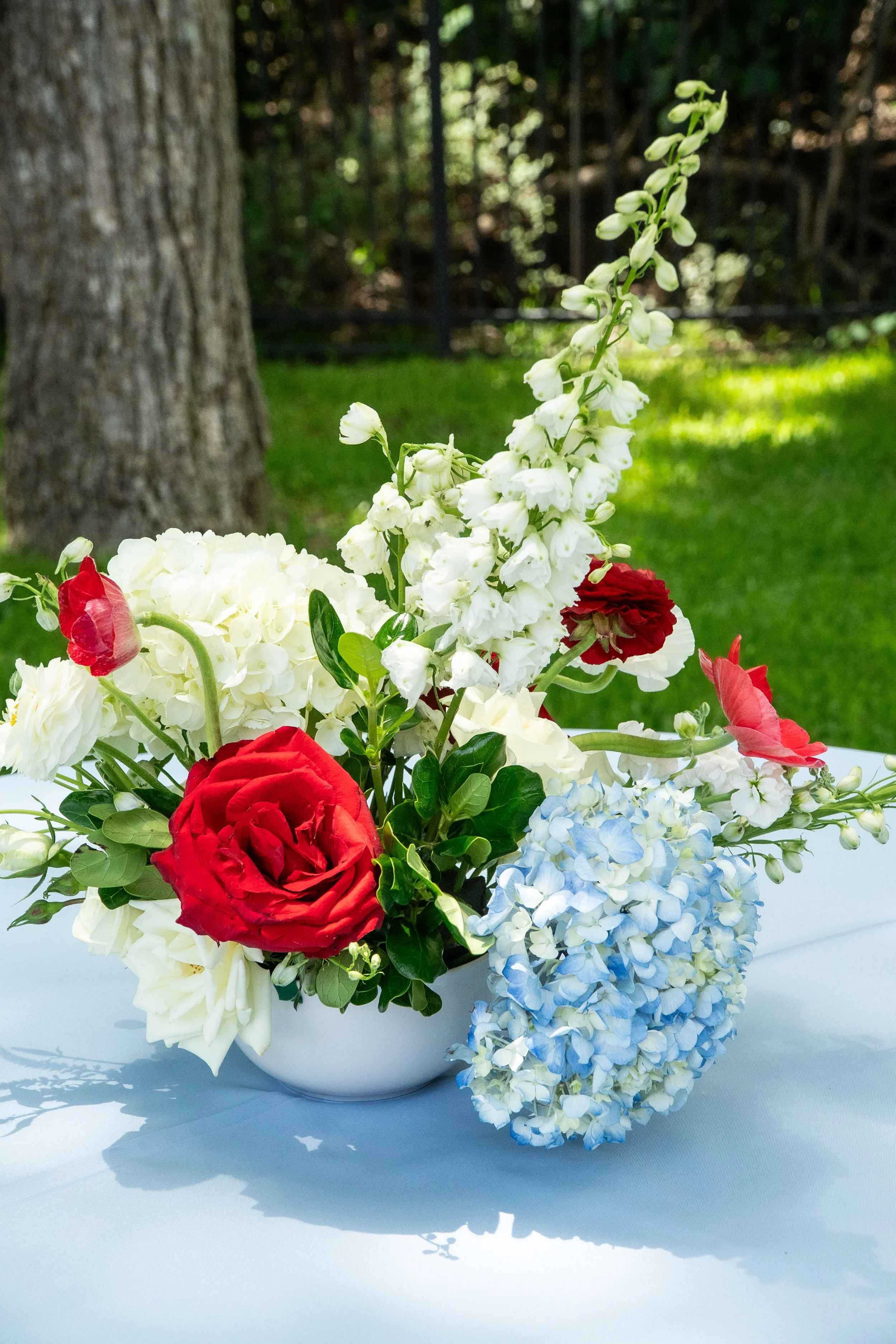 A floral arrangement in a white bowl with red, white, and light blue flowers, set outdoors on a white surface with a blurred tree and greenery in the background.