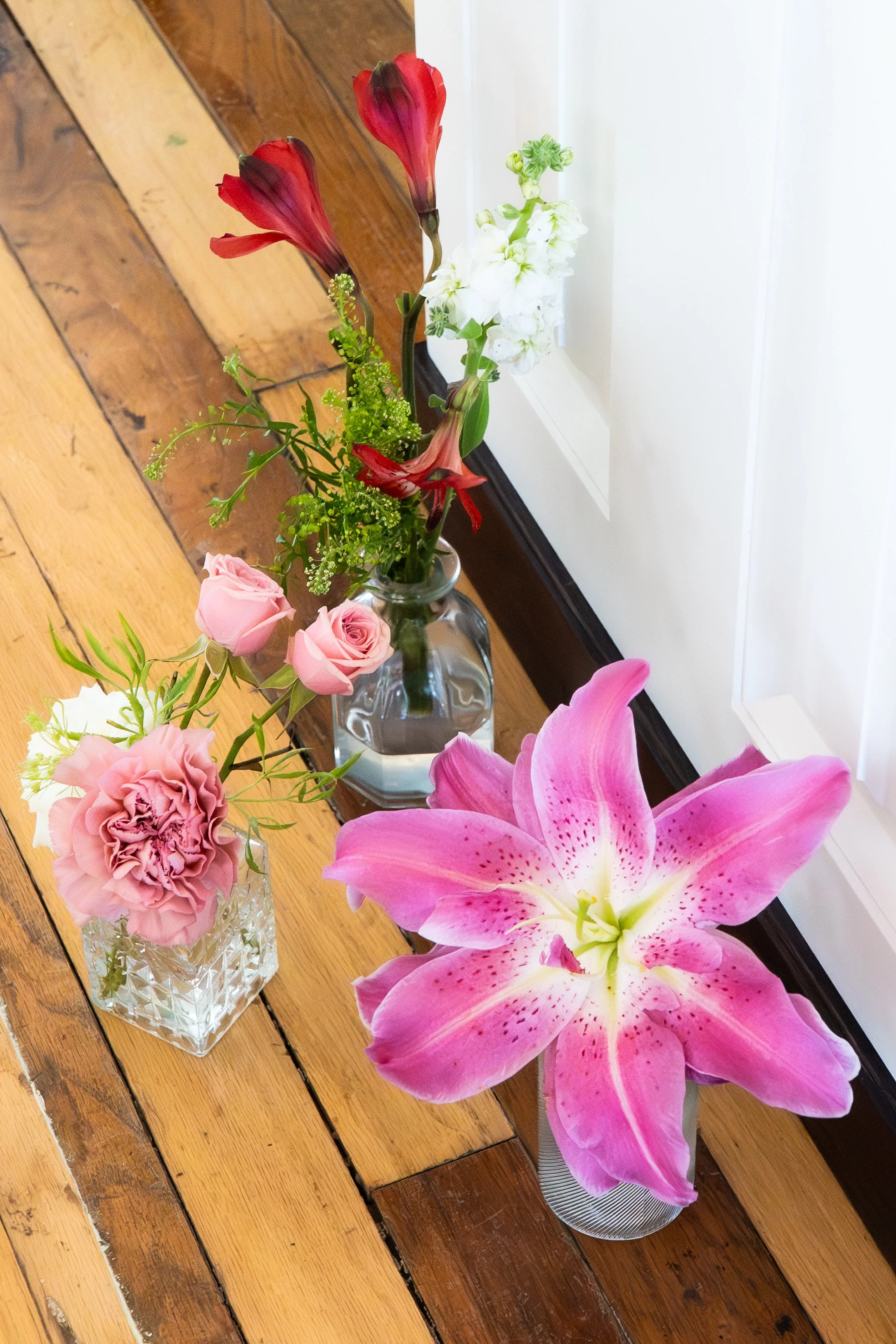 Three vases with pink, red, and white flowers resting on a wooden floor near a white door.