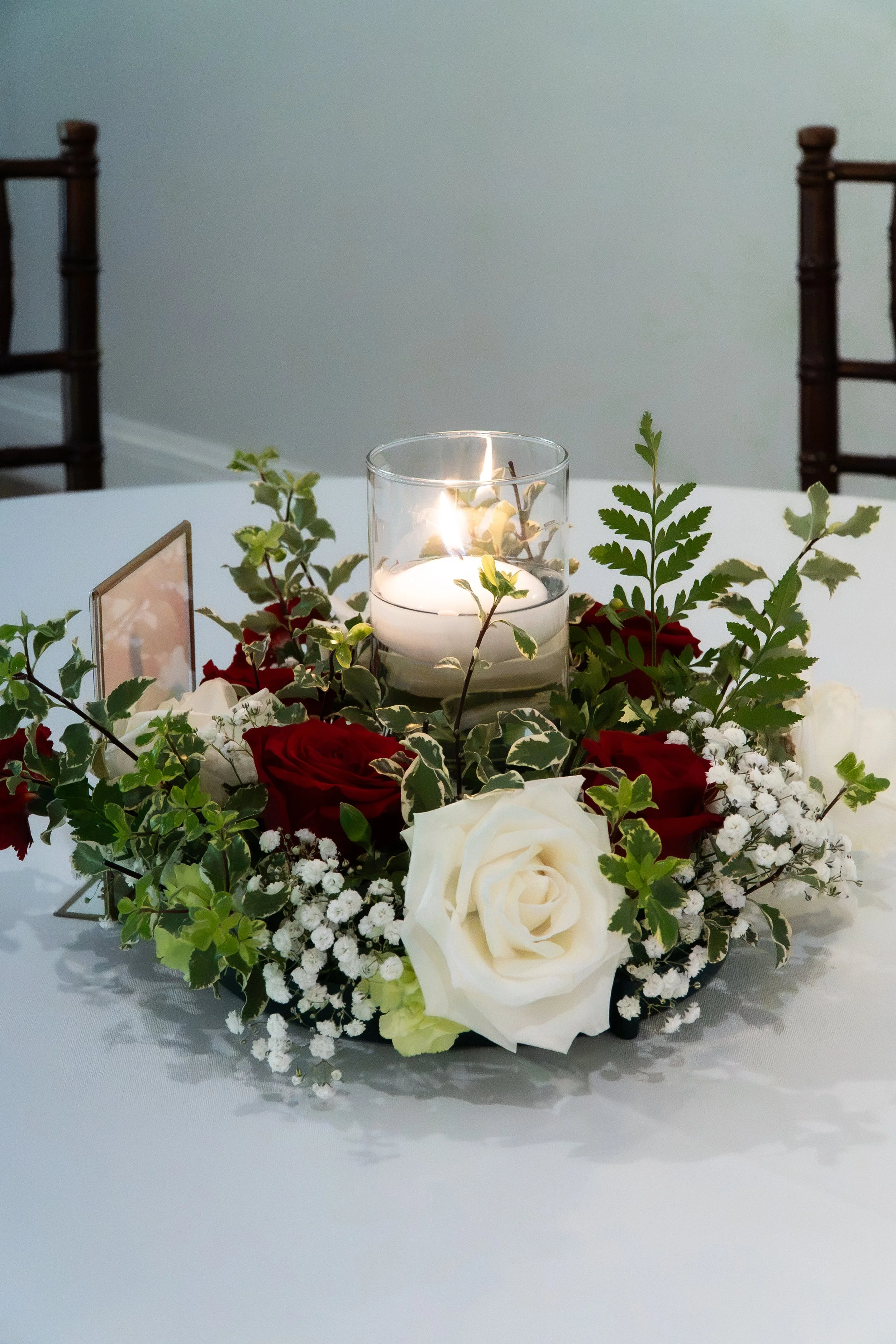 A floral arrangement with white roses, red roses, greenery, and baby's breath on a white table, with a glass candle holder and a small photo frame, in a softly lit room with light-colored walls.