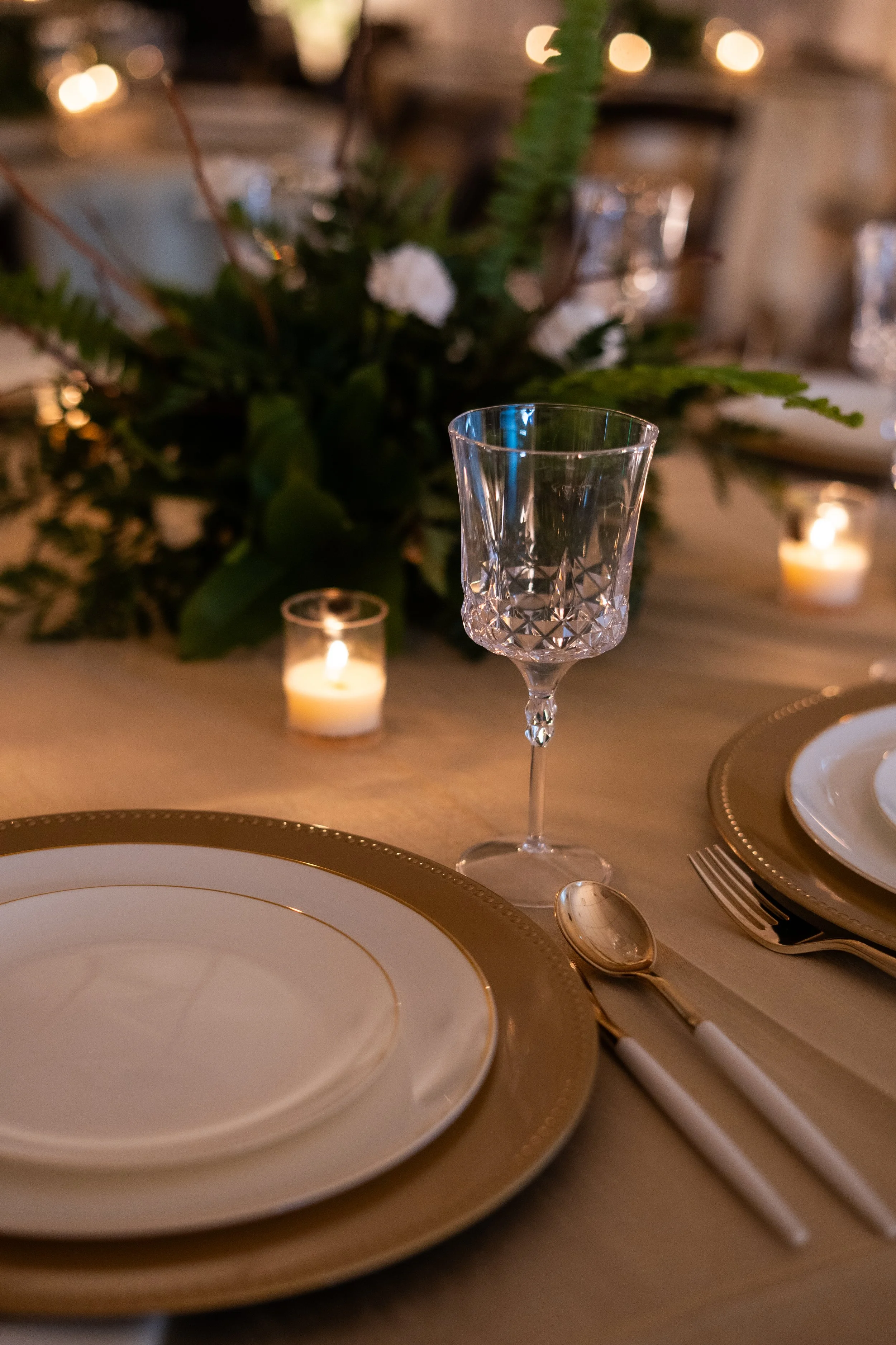 Elegant table setting with white plates with gold rims, silverware, a crystal wine glass, a candle, and a floral centerpiece with greenery and white flowers, creating a sophisticated ambiance.