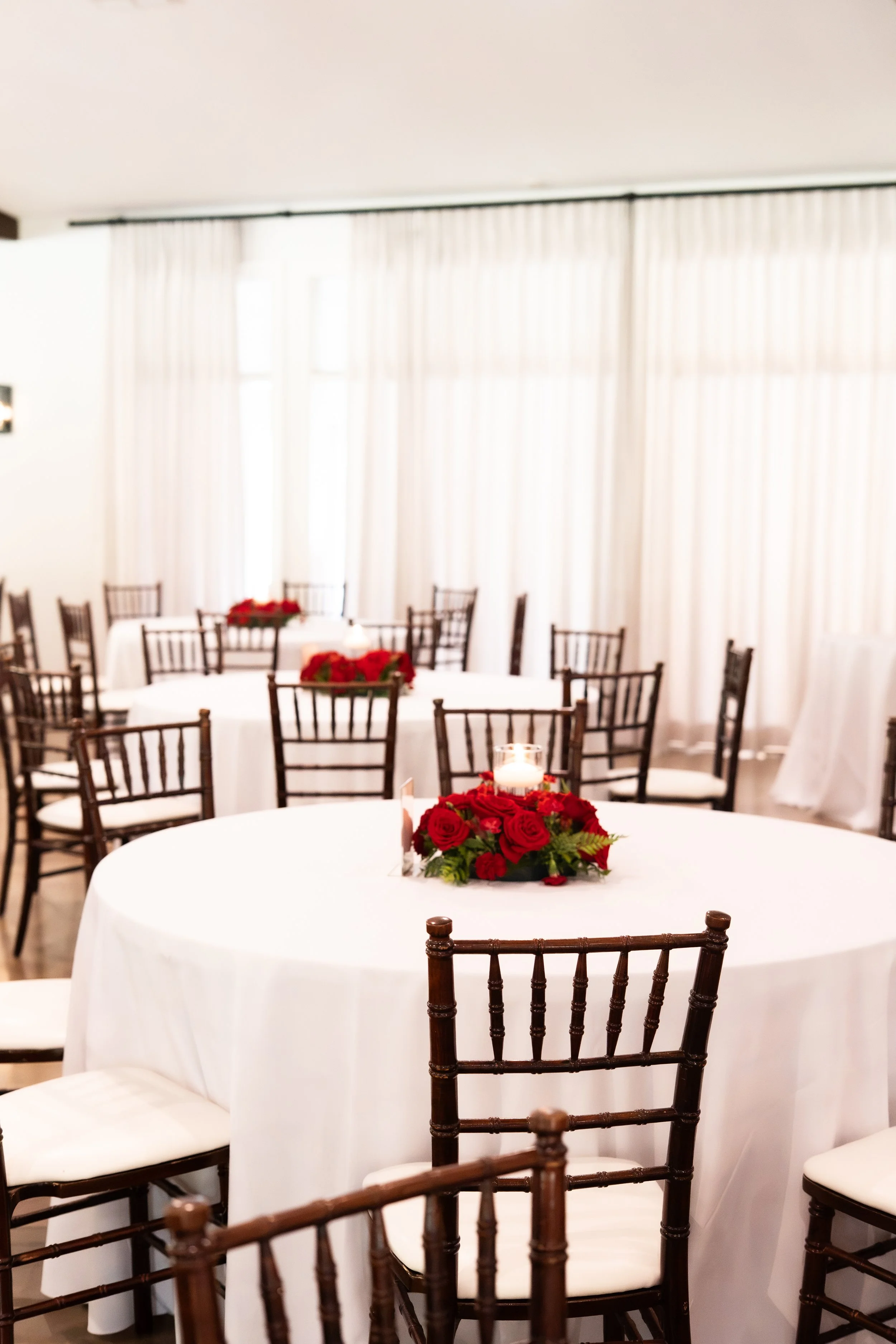 Elegant event room with round tables covered in white tablecloths, decorated with red floral centerpieces and surrounded by dark brown chairs with white cushions, near large windows with sheer curtains.