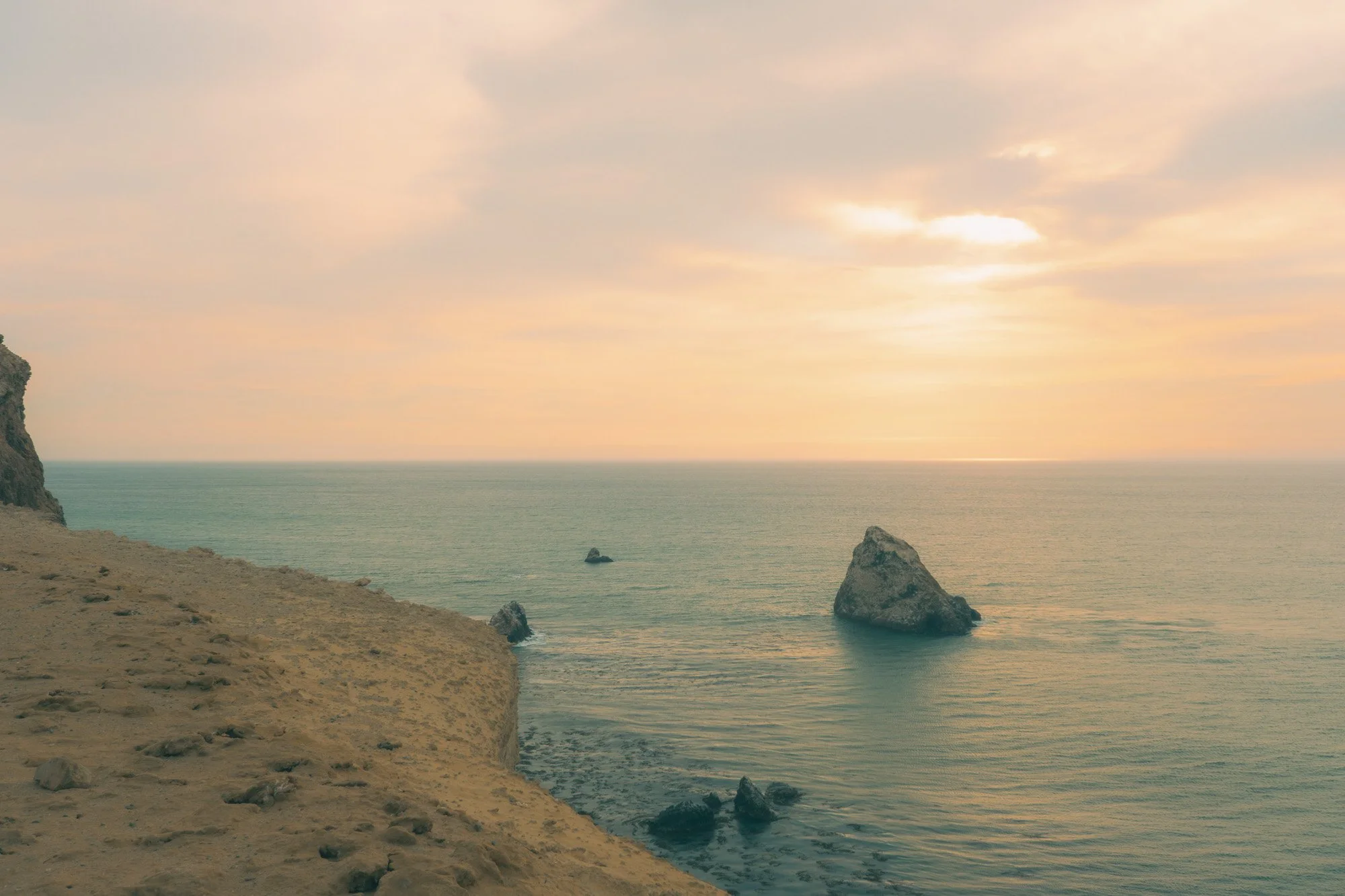 Sunset view of the ocean with large rocks near the shoreline and a cliff on the left side.