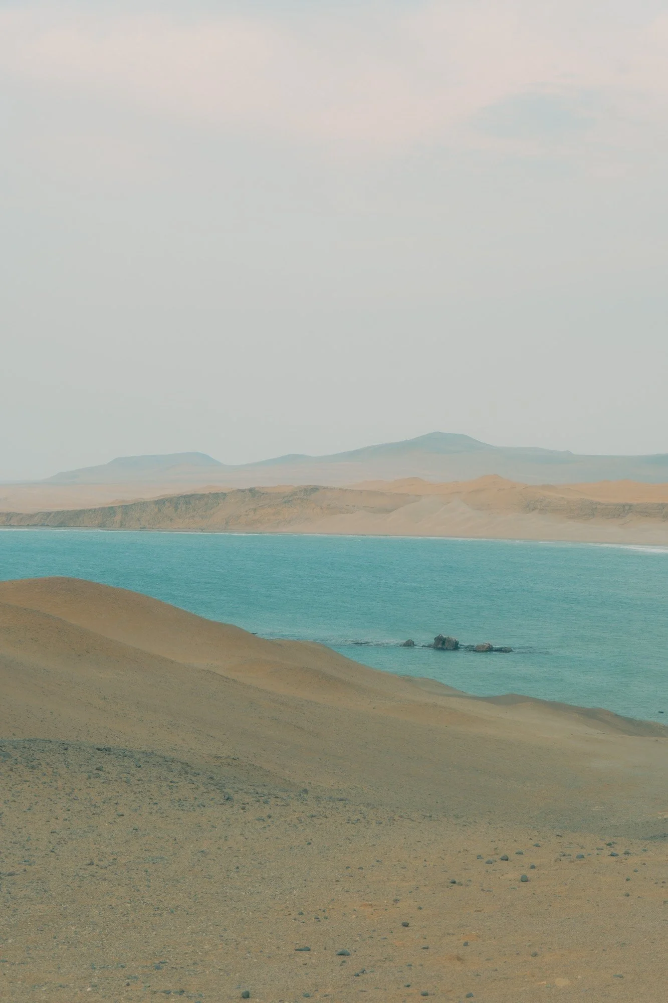 A desert landscape with sandy dunes and a body of water, possibly a lake or ocean, with mountains in the background and a cloudy sky.