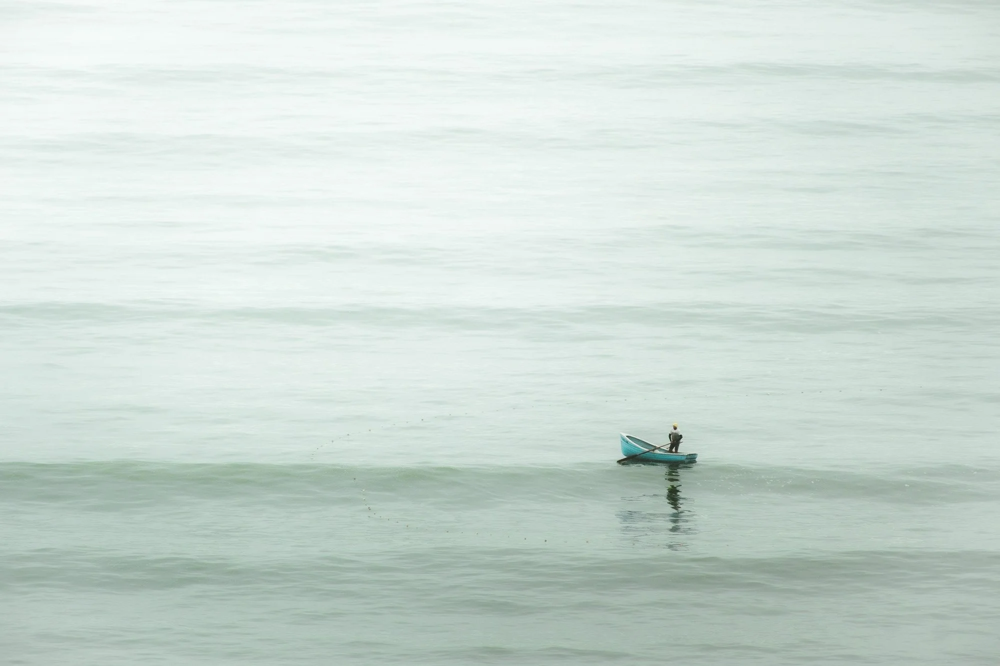 A person standing in a small blue boat on calm ocean waters with gentle waves.