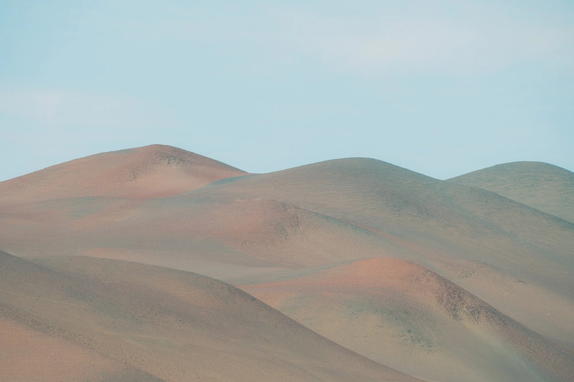 A landscape of rolling hills with pastel-colored soil and sparse vegetation under a light blue sky.