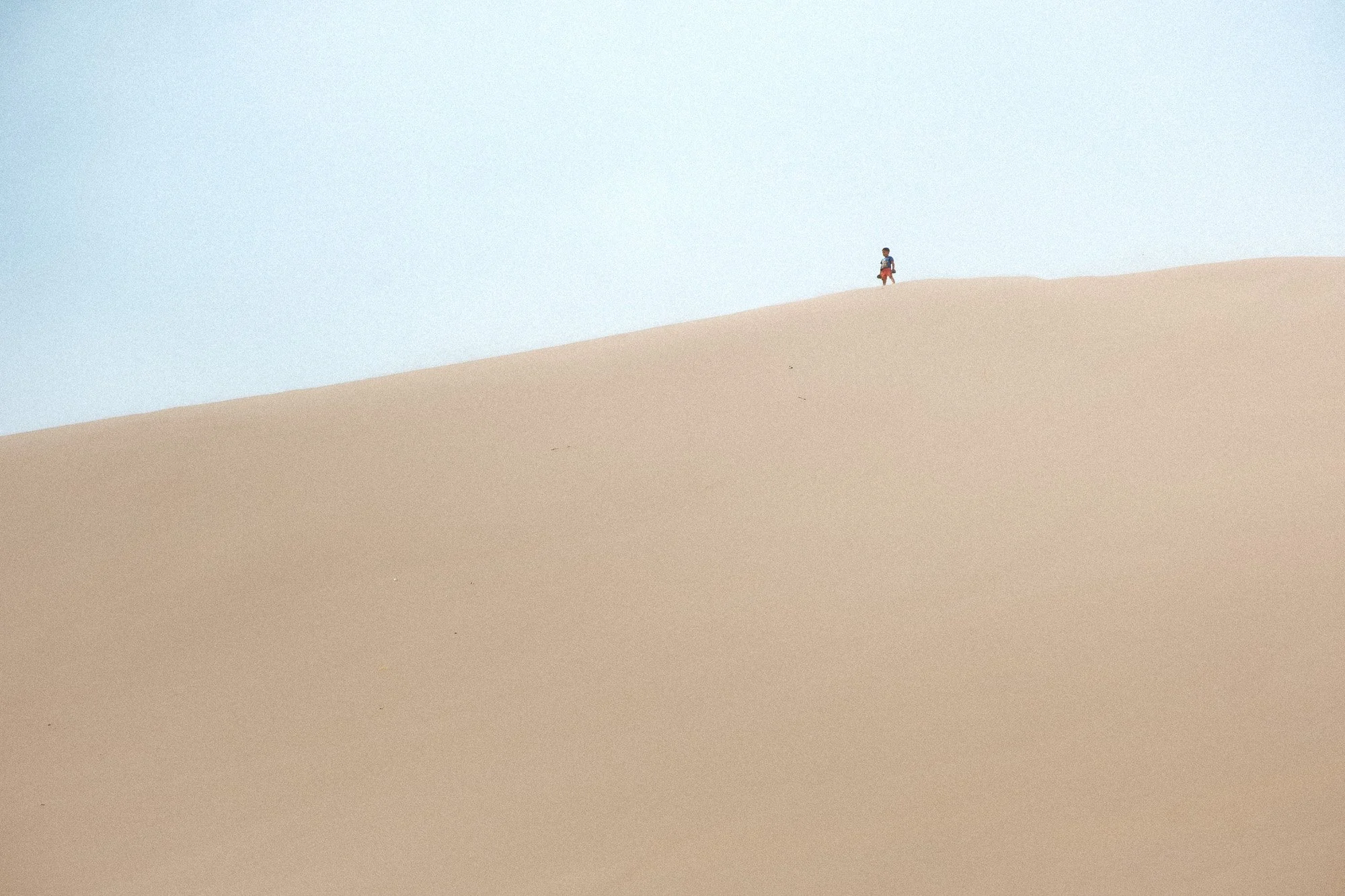 A small child standing alone on top of a sand dune with a vast, clear sky in the background.