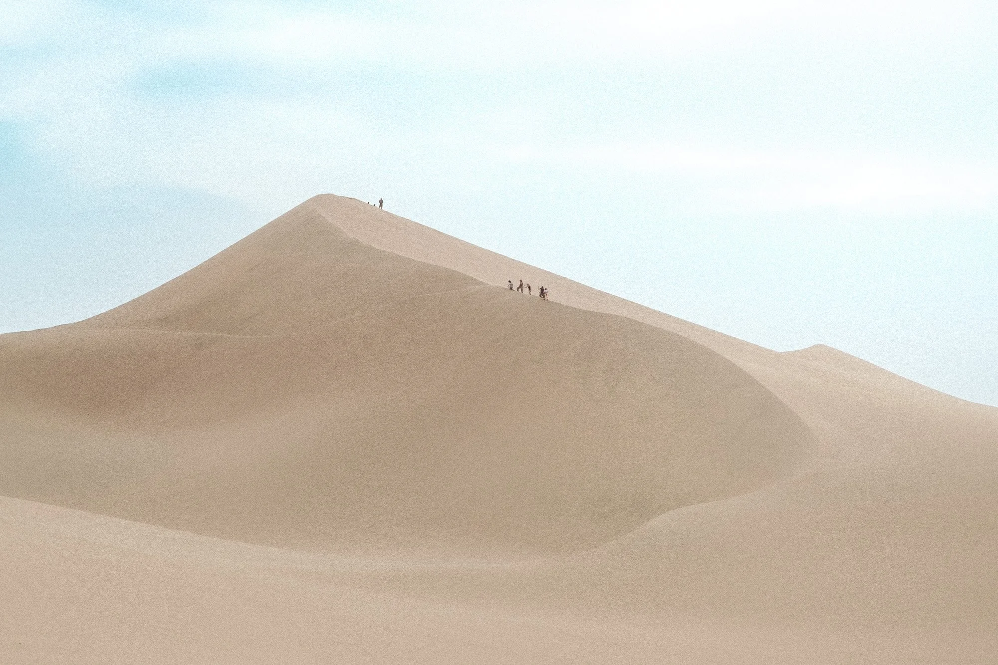 People walking on sand dunes in a desert landscape with a light blue sky.