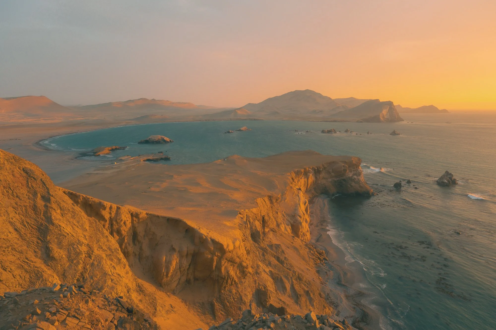 A coastal landscape at sunset with rugged cliffs, a sandy beach, a bay with water, and distant mountains.