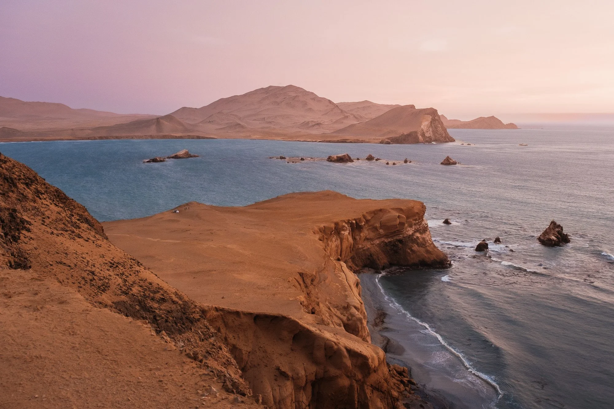 Scenic view of rocky cliffs overlooking the ocean with a distant mountain range and a pinkish sky at sunset.