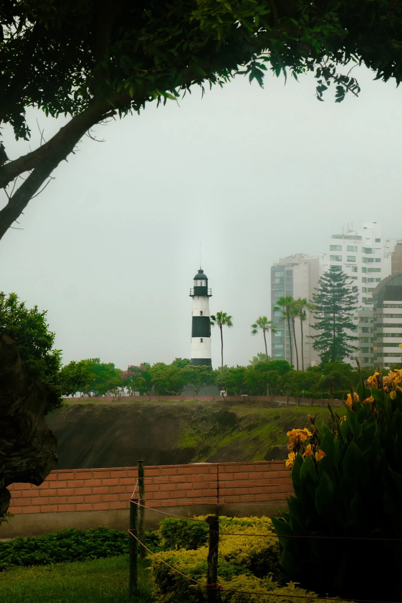 A lighthouse with black and white stripes situated on a green grassy area surrounded by trees with modern buildings in the background.