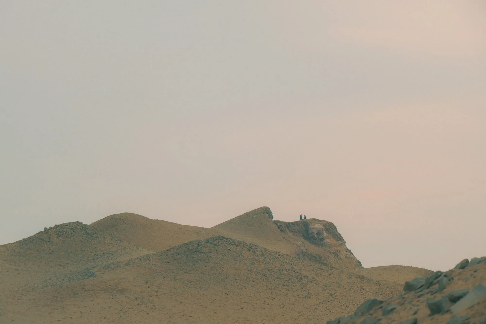 Desert landscape with sand dunes and a rocky formation in the distance, with two small figures standing on top of the tallest dune.
