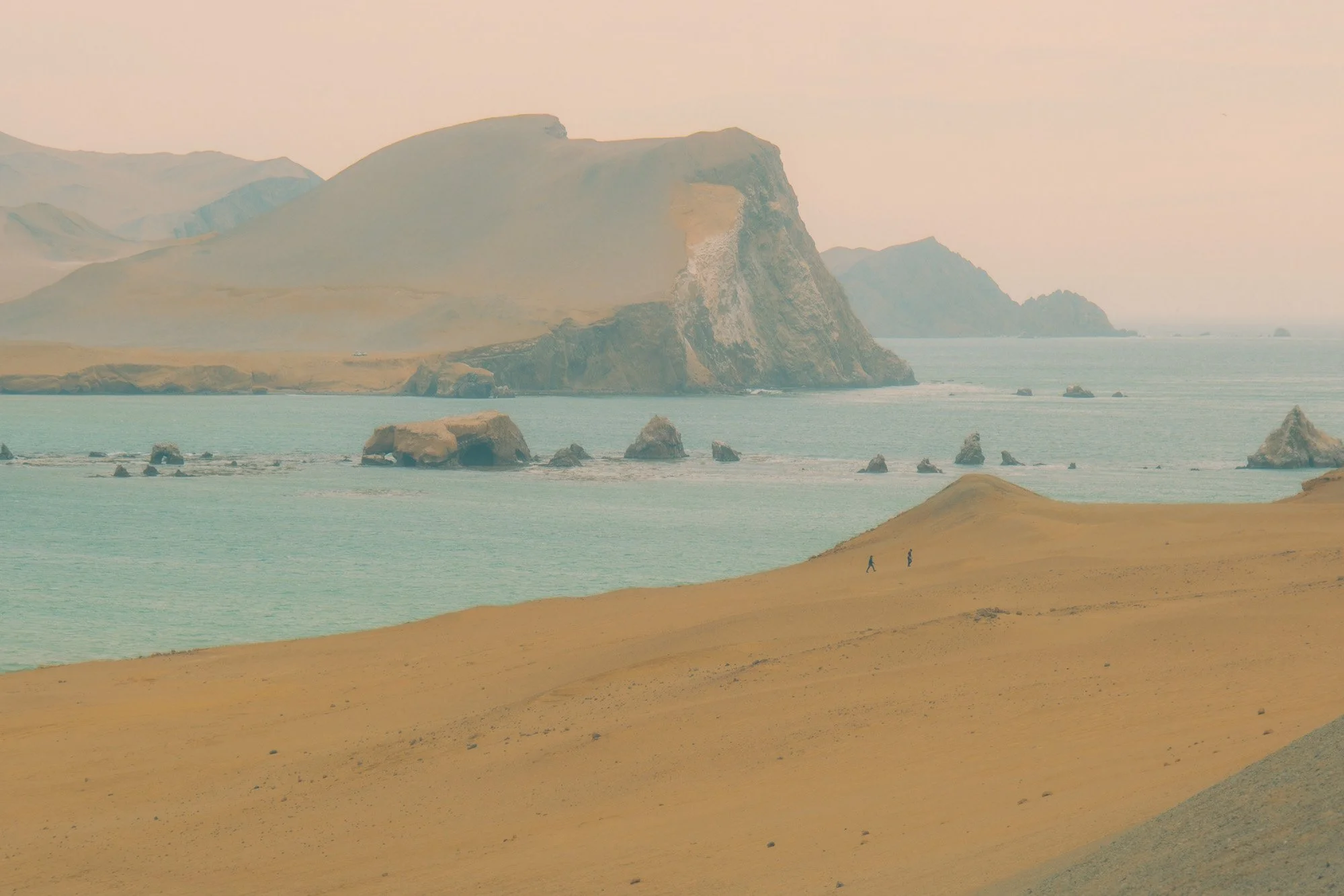 Landscape view of a coastal area with sandy dunes, turquoise water, rock formations, and large cliffs in the background under a soft pinkish sky.