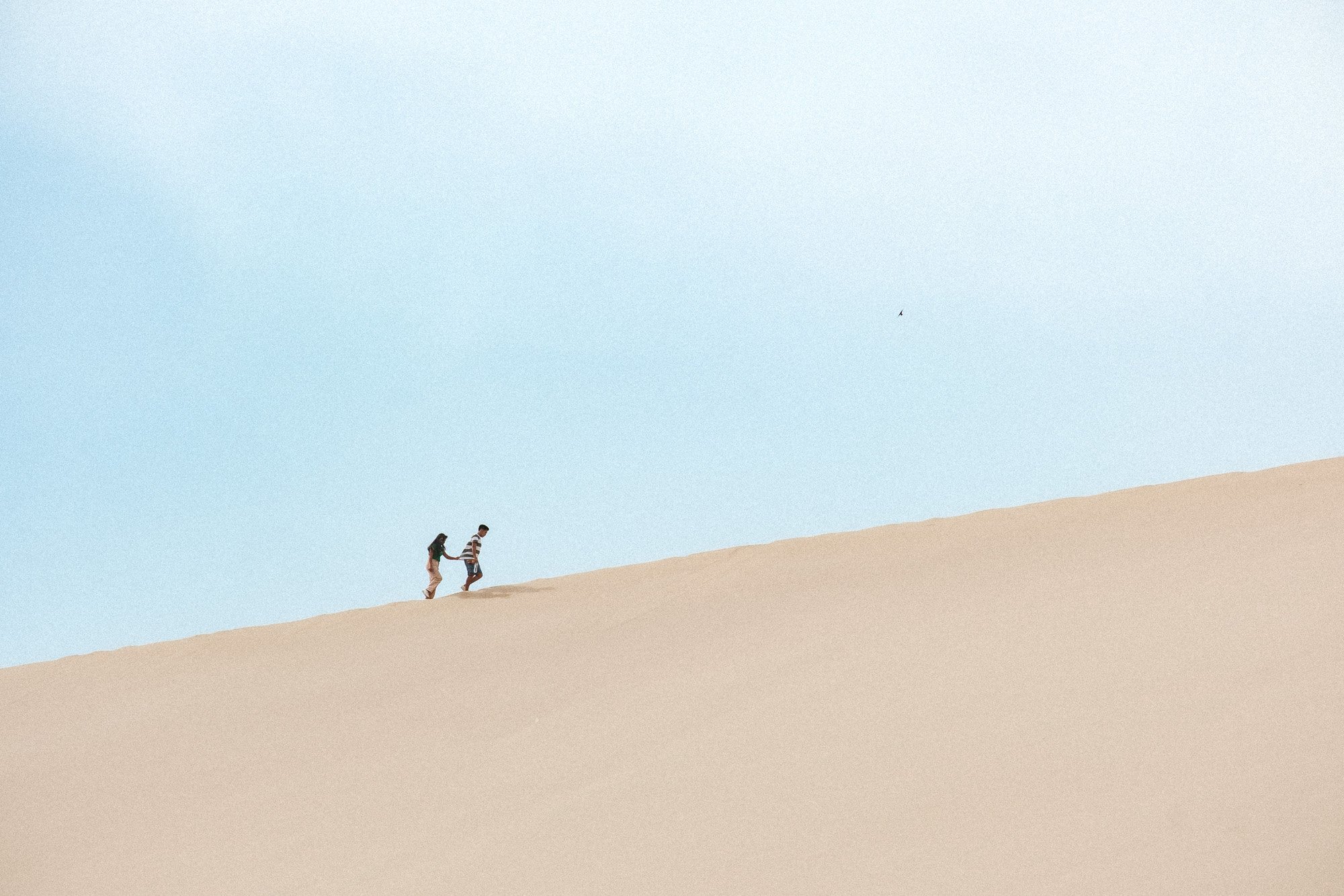 Two people walking up a sand dune in a desert with a clear blue sky.