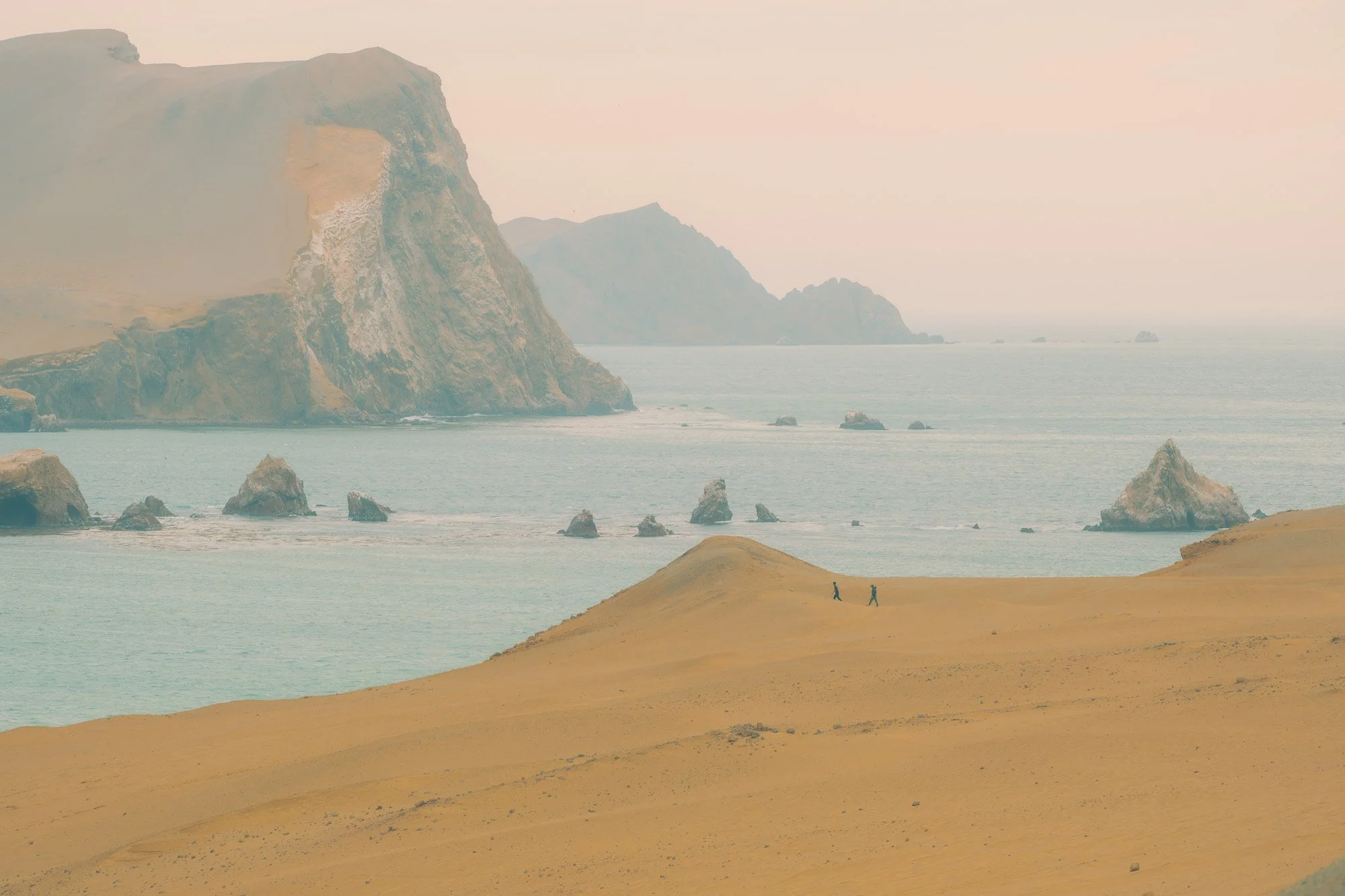 Two people walking on a sandy hill near the coast with large rocky cliffs and islands in the background under a pastel-colored sky.