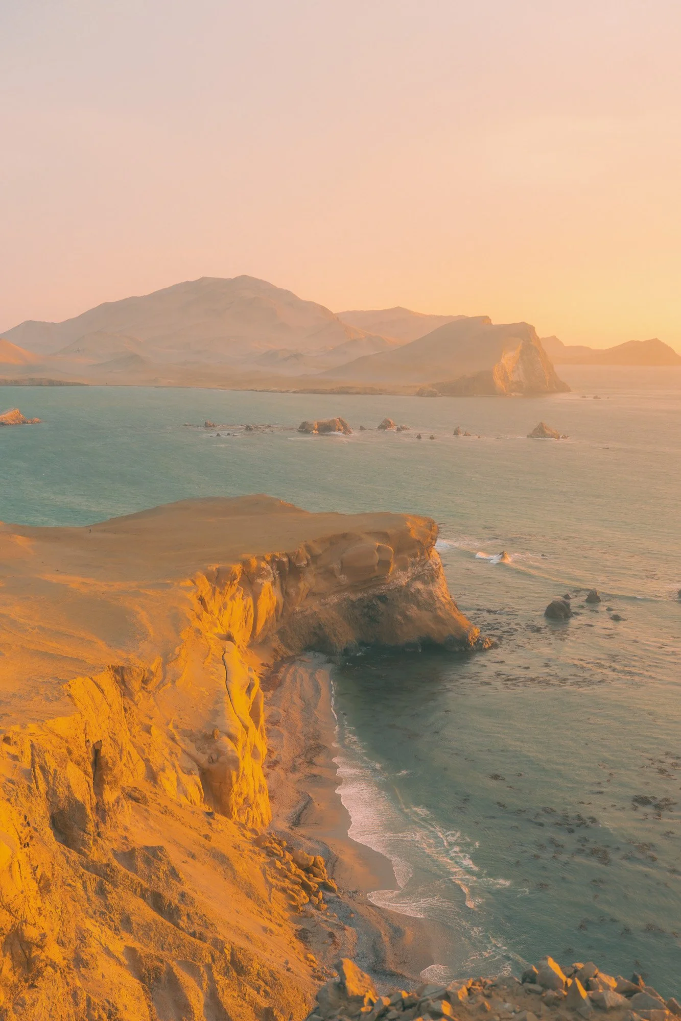 Sunset over rugged coastal cliffs with turquoise water, rock formations, and distant mountains in the background.