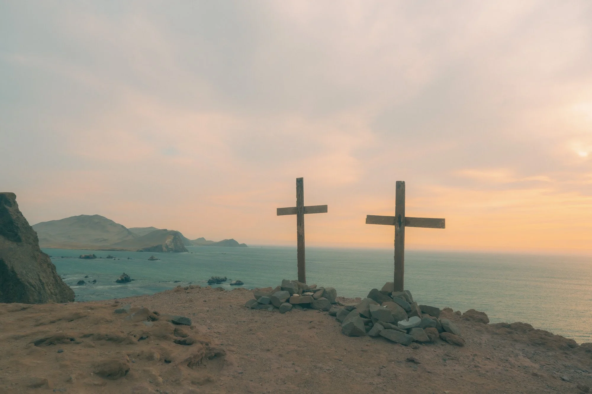 Two wooden crosses on a rocky hill overlooking the ocean at sunset or sunrise, with distant cliffs and a cloudy sky.