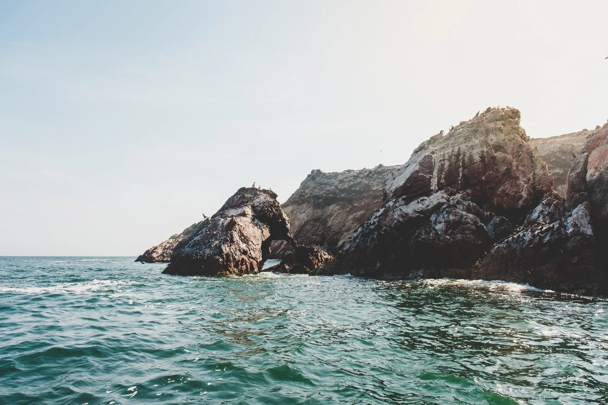 View of a rocky coastline with large rocks extending into the ocean under a clear sky.