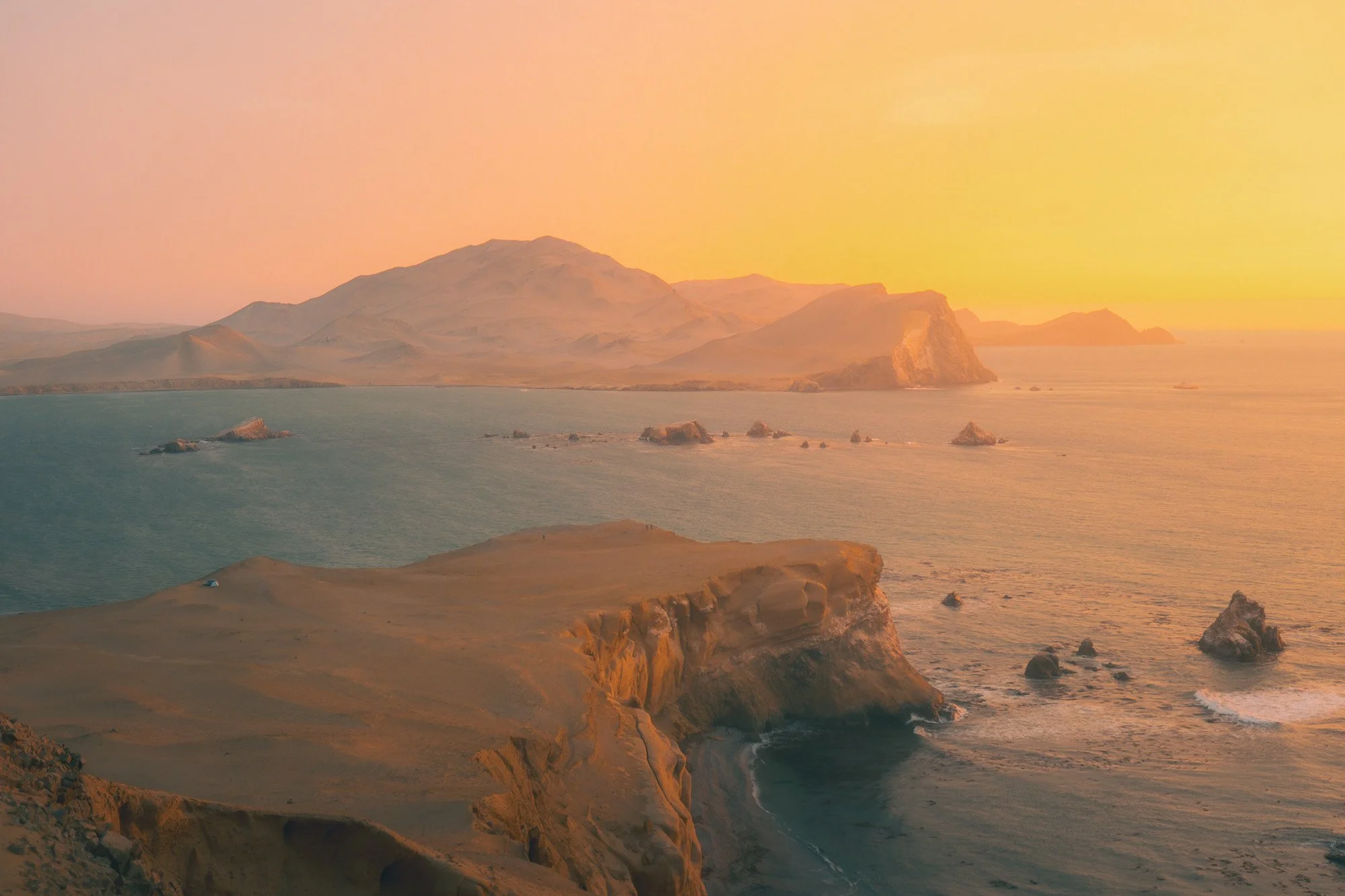 A scenic view of a coastline with cliffs, water, and distant mountains during sunset or sunrise.