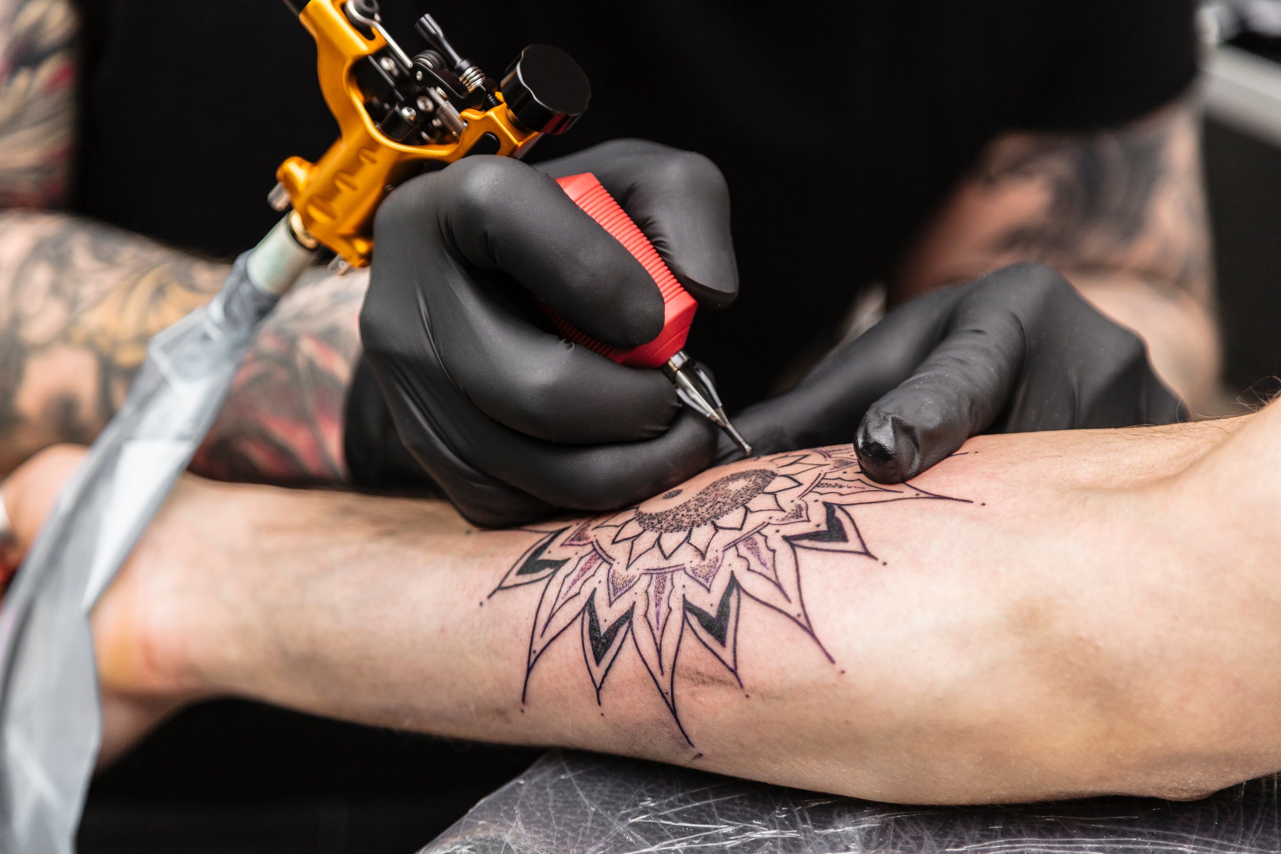 A tattoo artist in black gloves is creating a tattoo on a person's inner forearm, featuring a detailed sunflower design with black, pink, and purple ink.